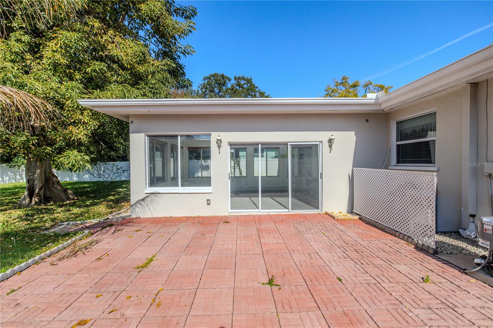 Spacious brick paver patio off the Florida room, offering easy indoor-outdoor living with sliding glass doors, mature shade trees, and a fenced backyard for privacy.