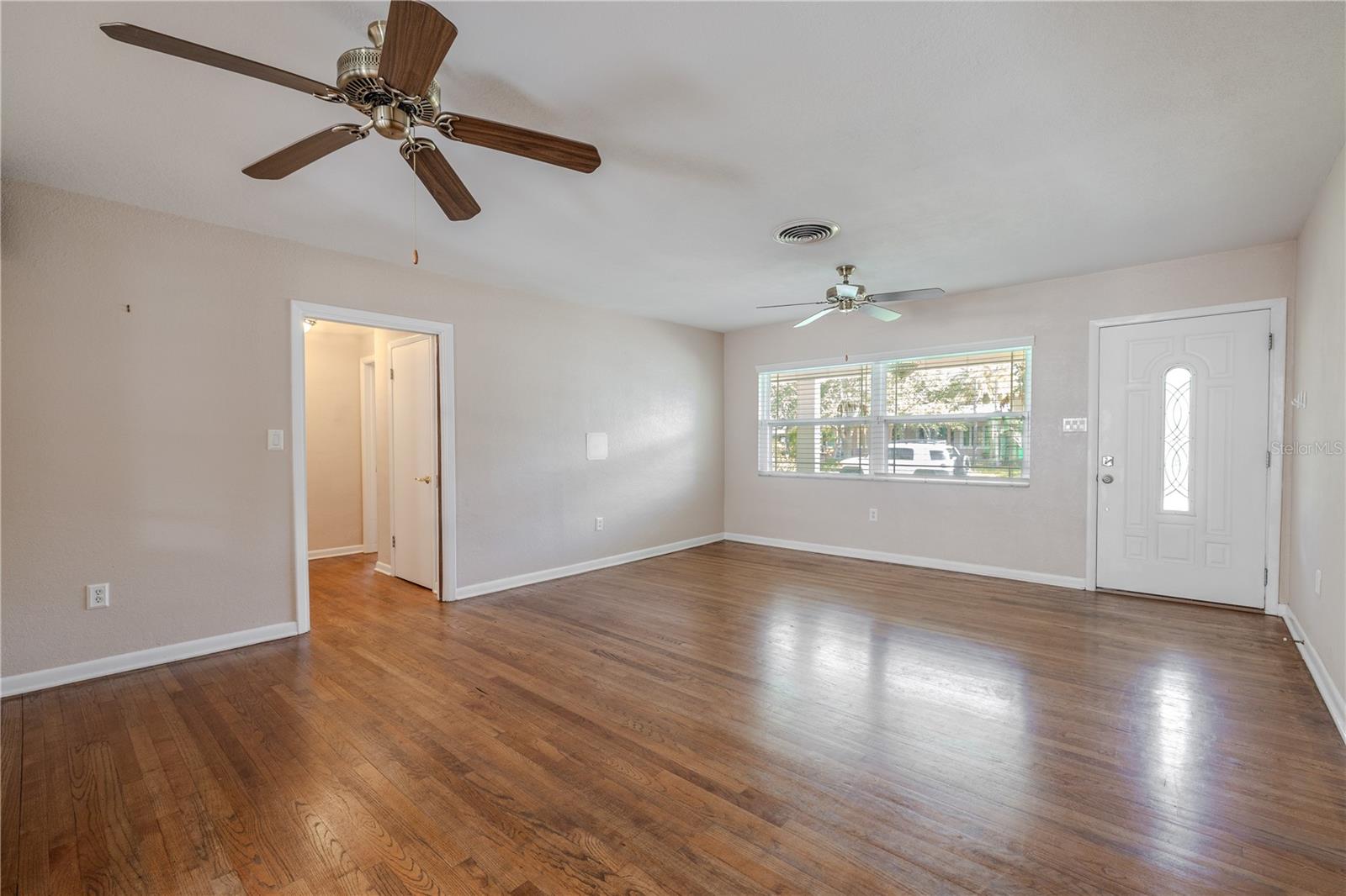 Additional view of the bright living room with beautiful hardwood floors, hallway access to the bedrooms/bathrooms, ceiling fans, and a large picture window that fills the space with natural light.