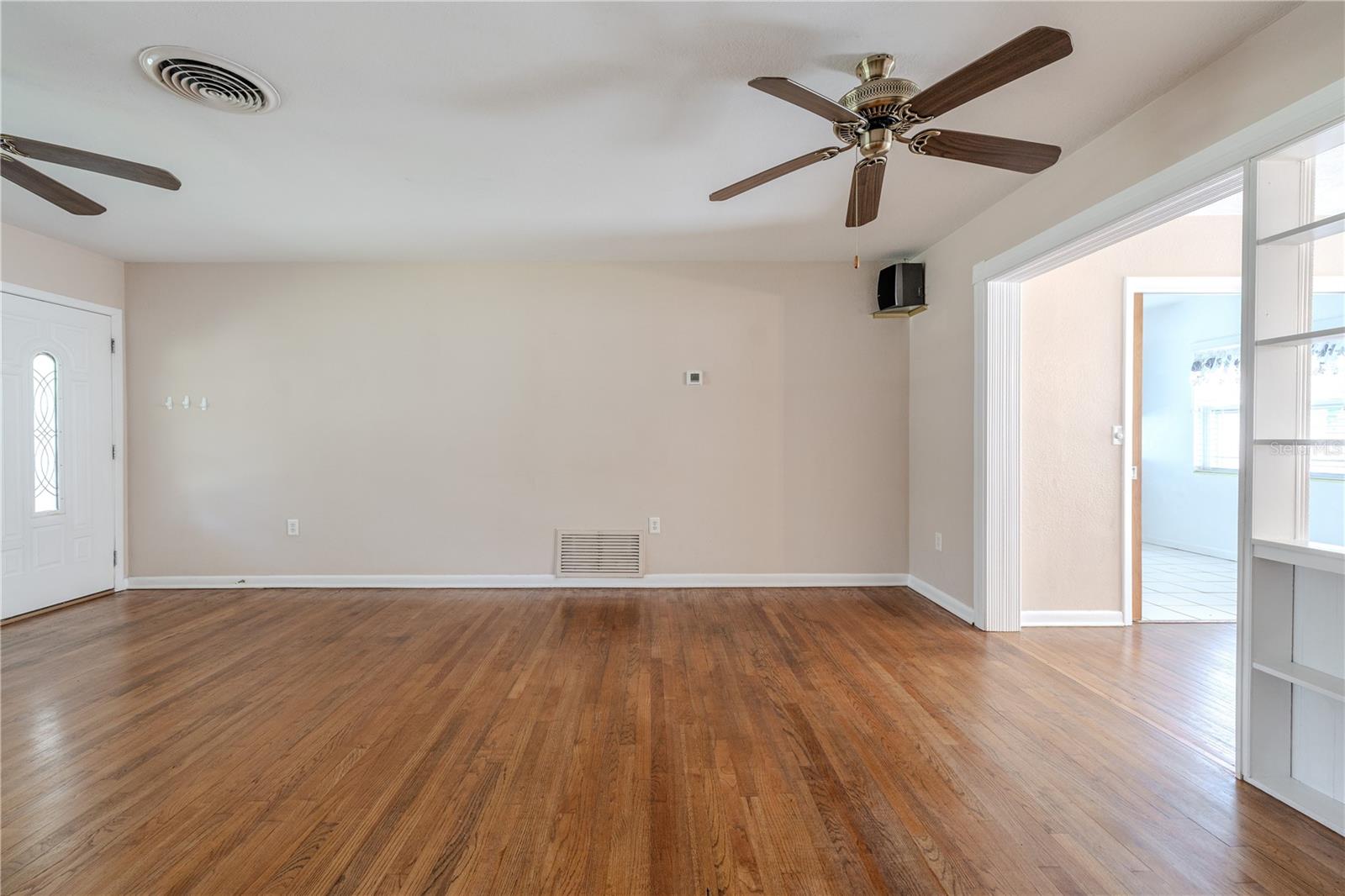 Large living room with gleaming hardwood floors and ceiling fans, offering an open connection to the adjacent dining room area with built-in shelving.