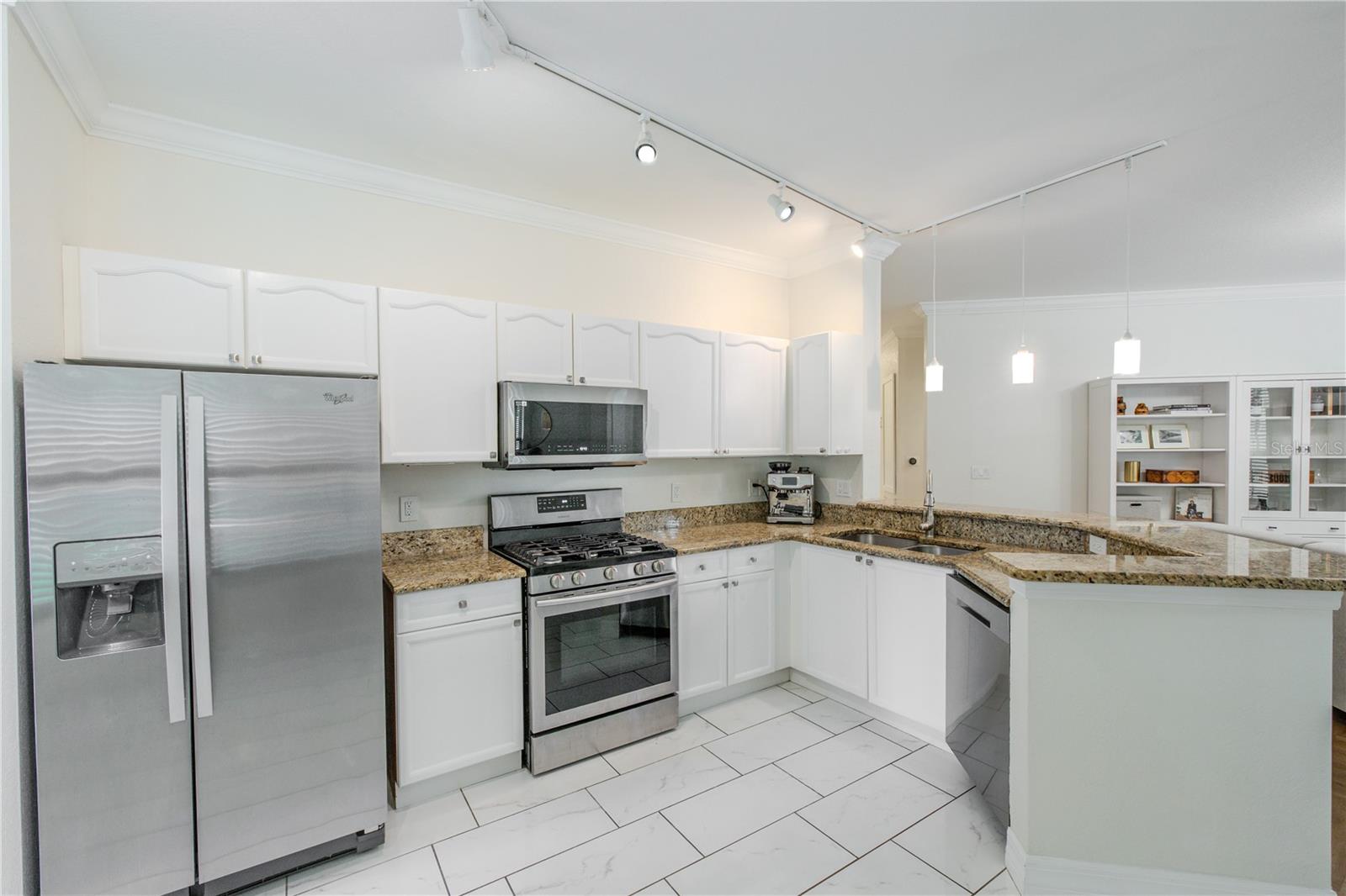 Stainless steel appliances—including a gas range, built-in microwave, refrigerator, and dishwasher—are thoughtfully arranged for efficiency. A double-basin sink with a gooseneck faucet overlooks the living area, reinforcing the kitchen’s open connection to the rest of the home.