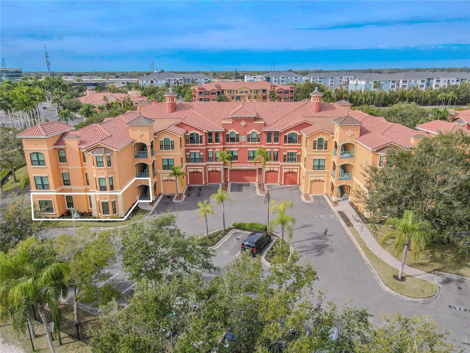 This aerial view highlights the striking Mediterranean-style architecture of Grand Bellagio at Baywatch, showcasing its warm stucco façade, decorative architectural accents, and classic barrel tile roofing. The building’s symmetrical design is anchored by elegant towers, arched openings, and covered breezeways that create a cohesive, resort-inspired aesthetic.