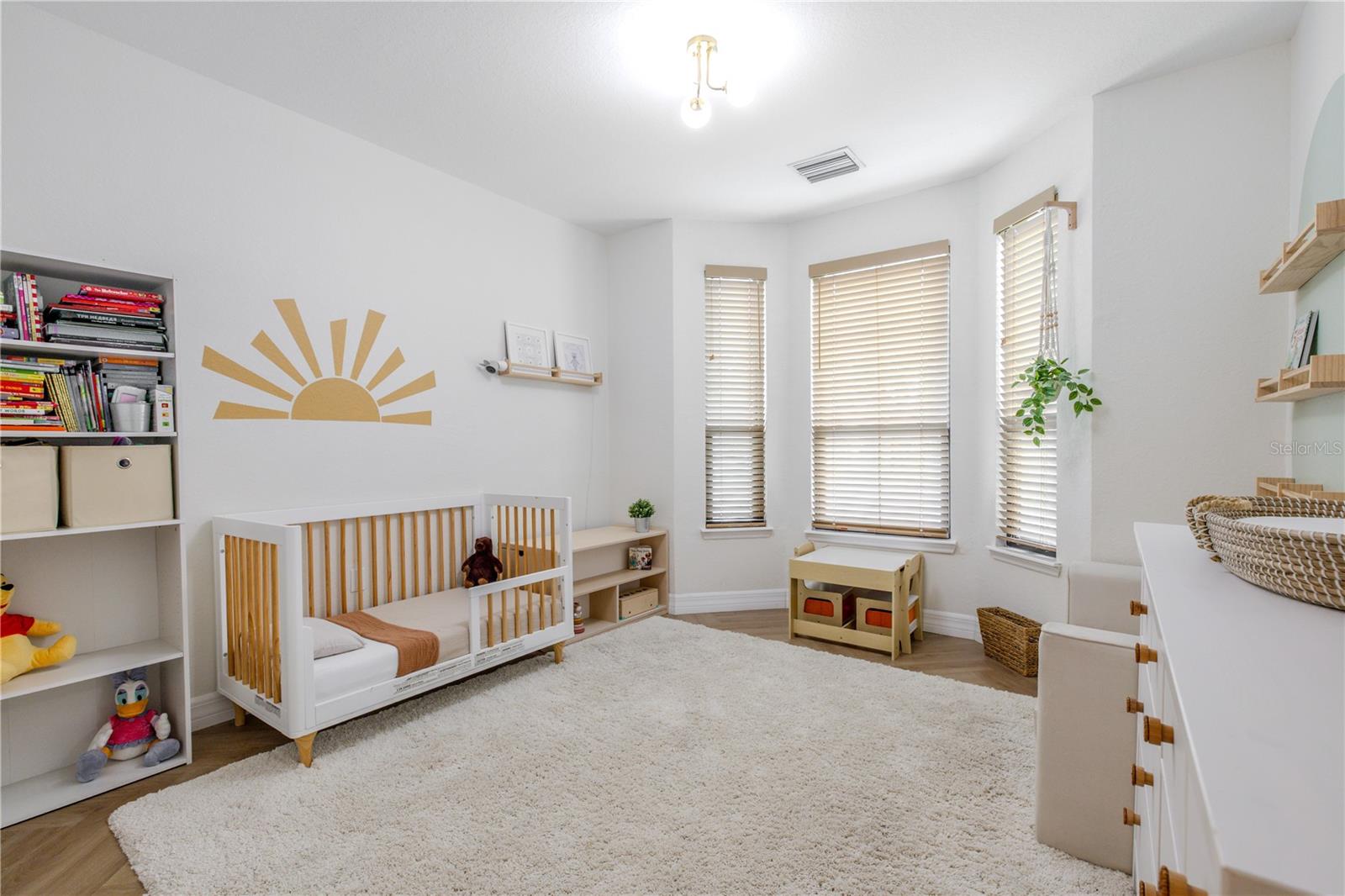 Bedroom 2 is bright, airy, and thoughtfully styled, featuring soft neutral walls, warm wood-look luxury vinyl flooring, and abundant natural light from multiple windows fitted with classic wood blinds. The spacious layout easily accommodates a nursery, guest room, or home office, with room to grow and reconfigure over time.