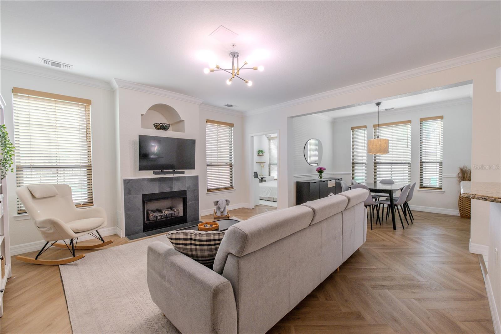 The living room has a bright, open layout with wide-plank flooring laid in a subtle herringbone pattern adding warmth and visual interest, while crisply painted walls and crown molding create a clean, timeless backdrop. Natural light filters in through multiple windows fitted with wood-look blinds, giving the space an airy, well-balanced feel throughout the day.