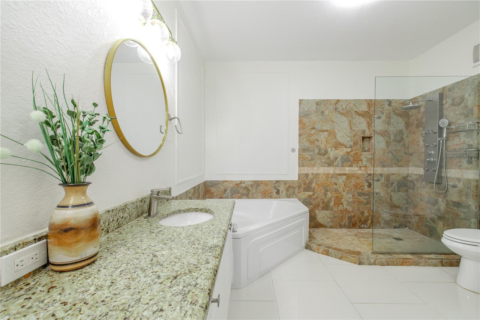 The primary ensuite bathroom is designed with a spa-inspired layout that includes a granite-topped vanity with undermount sink, crispcabinetry, and elegant lighting paired with a framed mirror. A soaking tub anchors the space, while the walk-in glass-enclosed shower features natural stone tile, a built-in niche, and a multi-function shower system for a true retreat experience.