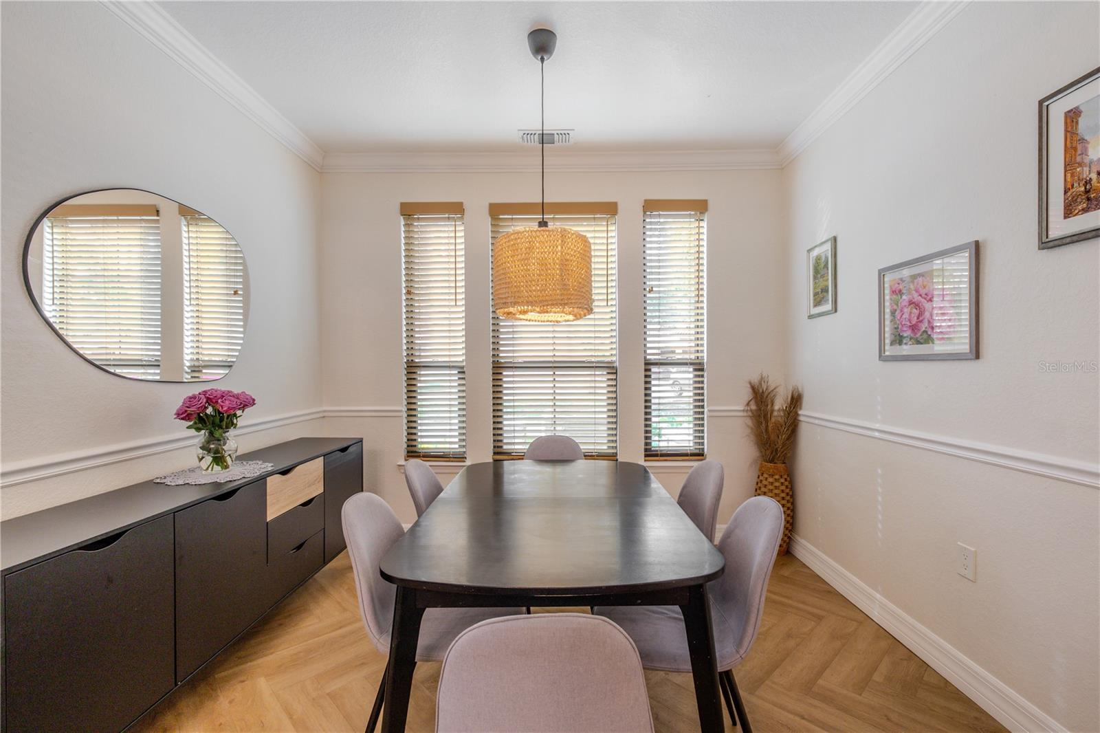 The dining room is bright and well-proportioned, defined by clean architectural lines and a calm, inviting atmosphere. Three tall windows fitted with wood-look blinds bring in abundant natural light, while crisp white walls and crown molding add a timeless finish.