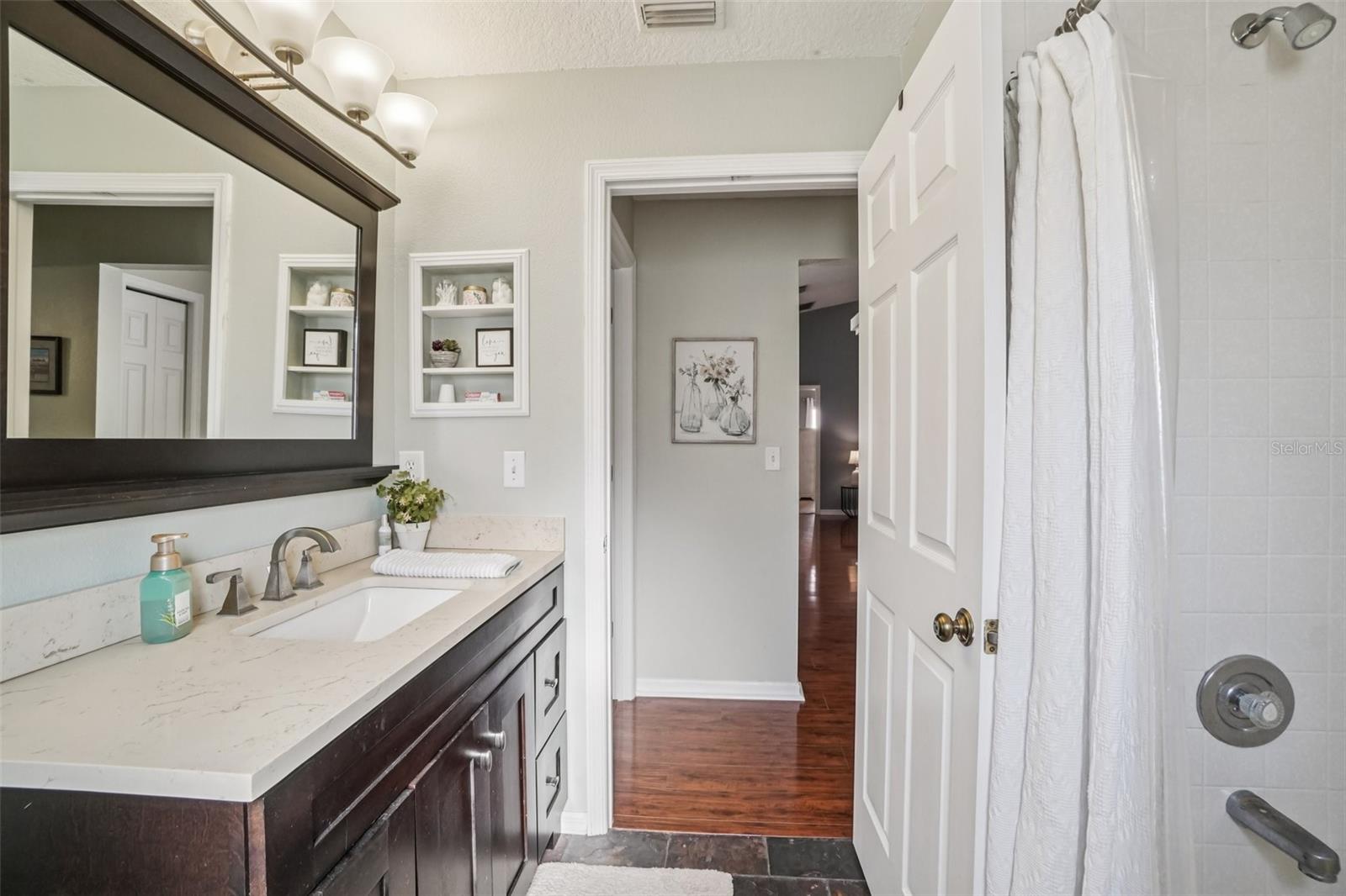 Bathroom 1 – Vanity ViewUpdated bathroom featuring a dark wood vanity with stone countertop, undermount sink, and modern fixtures, accented by a framed mirror and built-in wall niches for added storage.