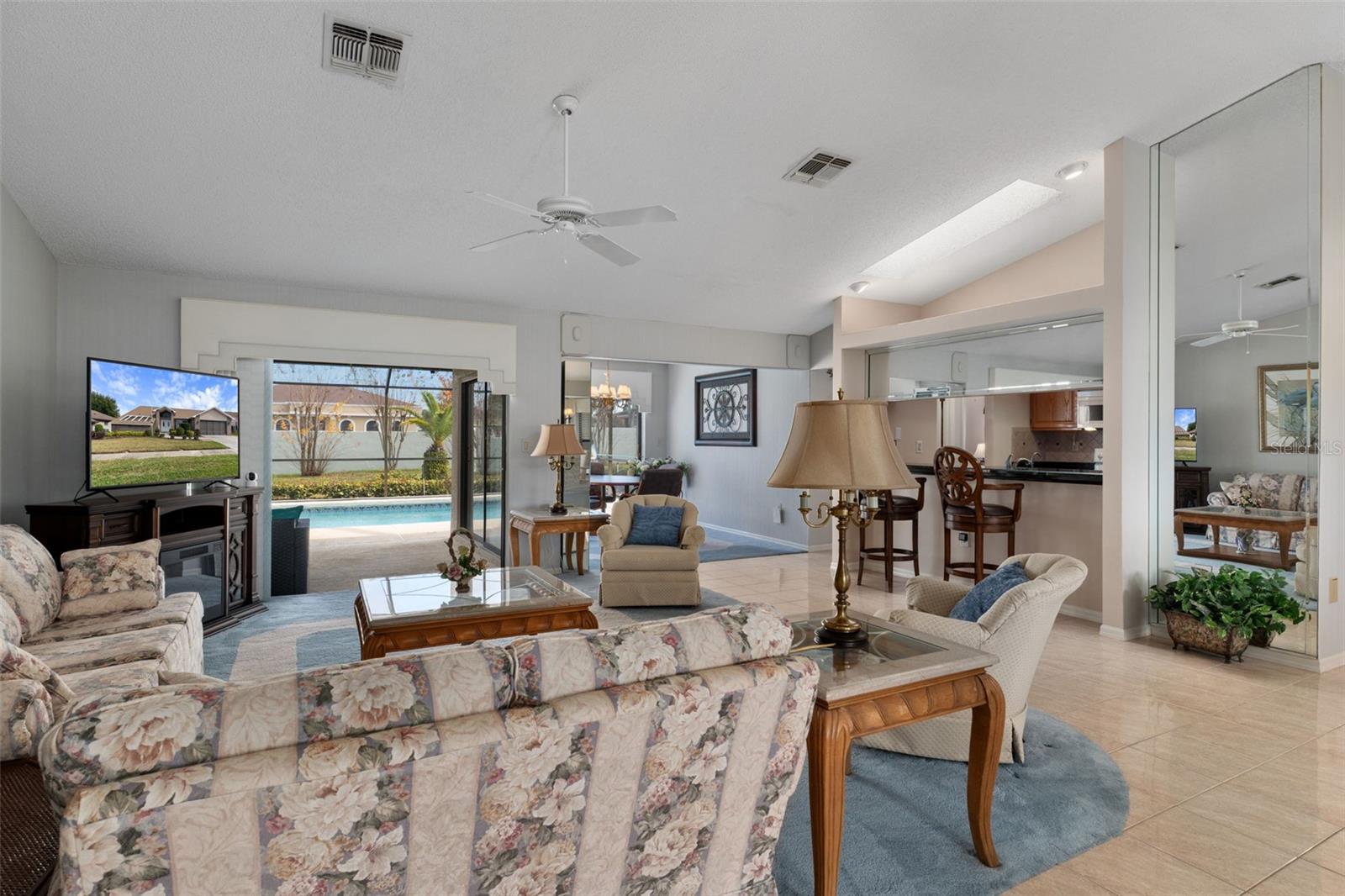 This spacious great room with high ceilings is open & bright.  The breakfast nook in the back also overlooks the pool/patio.  The kitchen w/ bar seating, a skylight and plant shelf overhead is to the right.