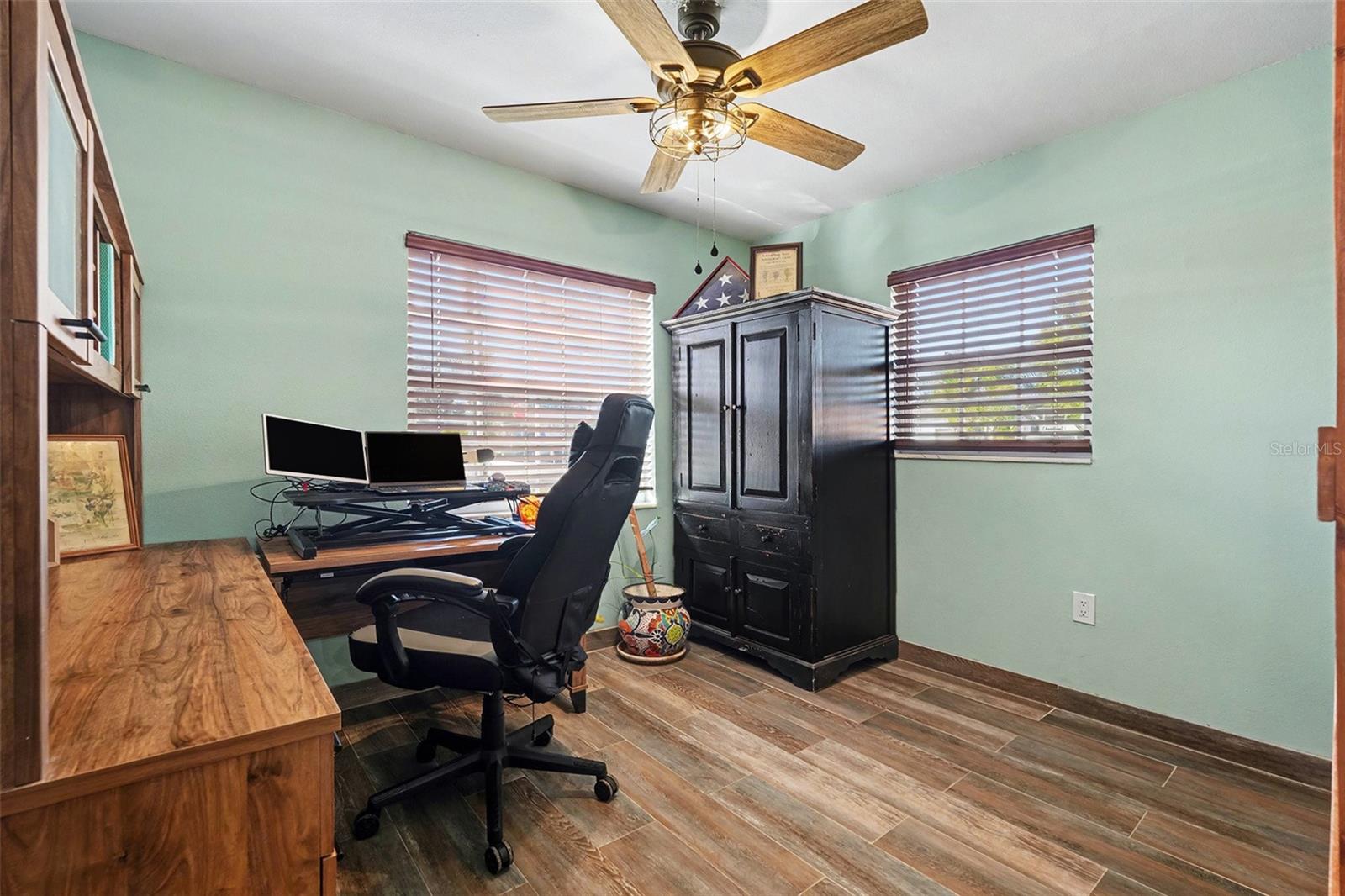 Dedicated home office or flex space featuring wood-look tile flooring, ceiling fan, and dual windows providing abundant natural light—ideal for remote work or study