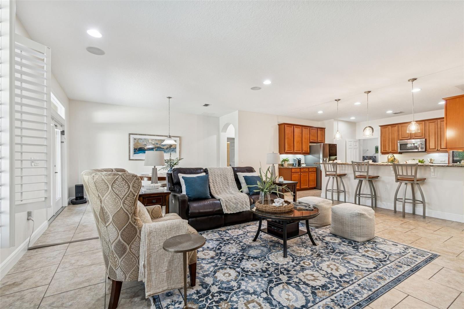 The inviting family room just off the kitchen, featuring an open, light-filled layout with tile flooring, large windows with plantation shutters, and ample space for relaxing, gathering, and entertaining—perfectly positioned for everyday living and effortless connection to the heart of the home.