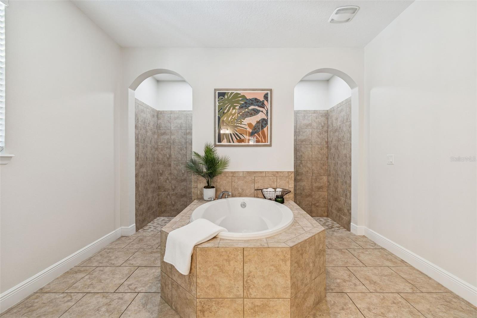 A bright, spa-like primary bathroom featuring a raised tiled soaking tub, symmetrical arched alcoves that lead to walk-in shower, warm neutral finishes, and clean & elegant