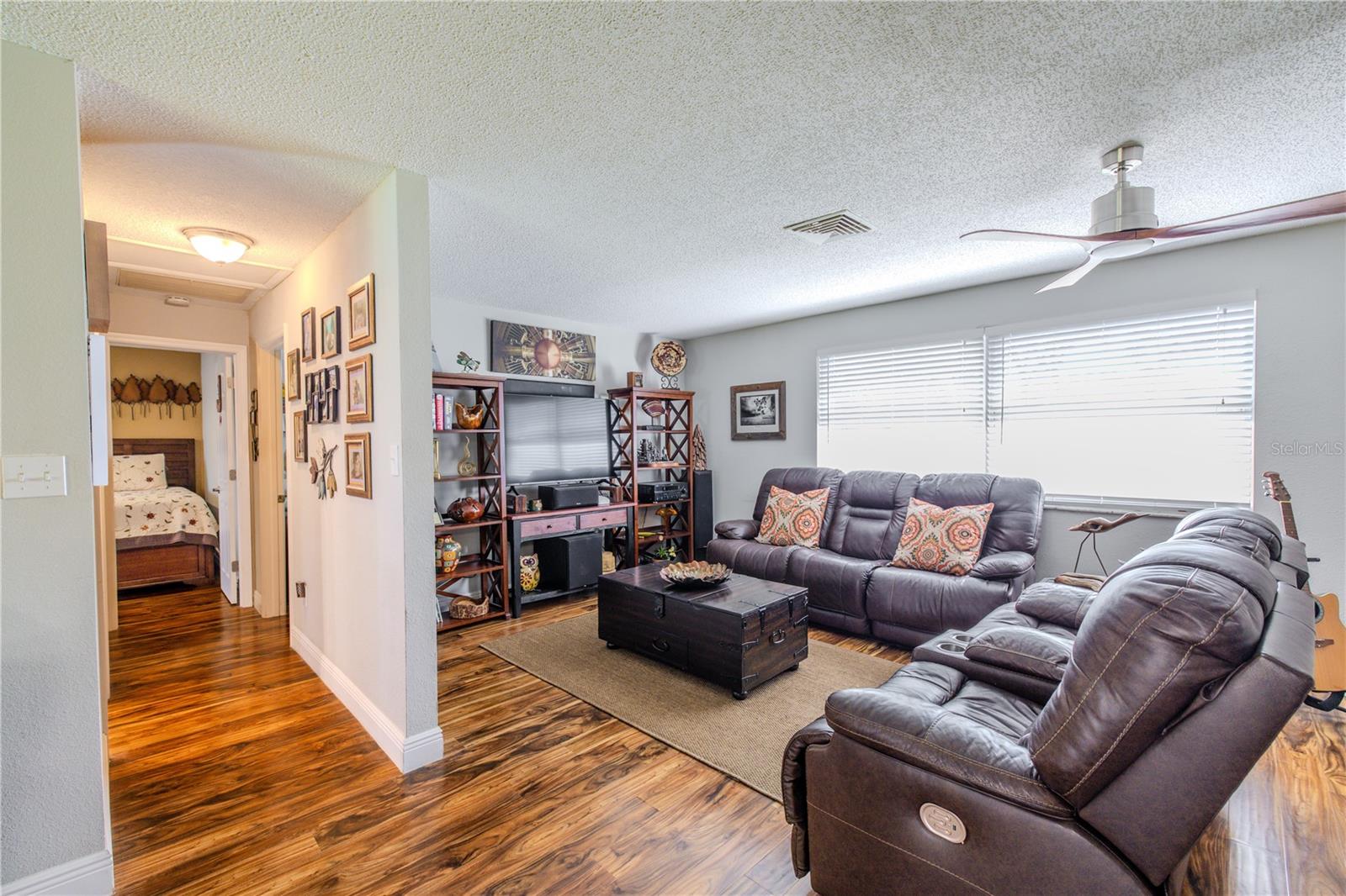 Open-concept layout connecting the kitchen, dining, and living areas for a cohesive and comfortable flow. The living room features wide windows with blinds, ceiling fan, and laminate flooring, while the adjacent kitchen showcases granite counters, stainless steel appliances, and ample cabinetry. A central hallway provides separation between living spaces and bedrooms without interrupting the open feel.