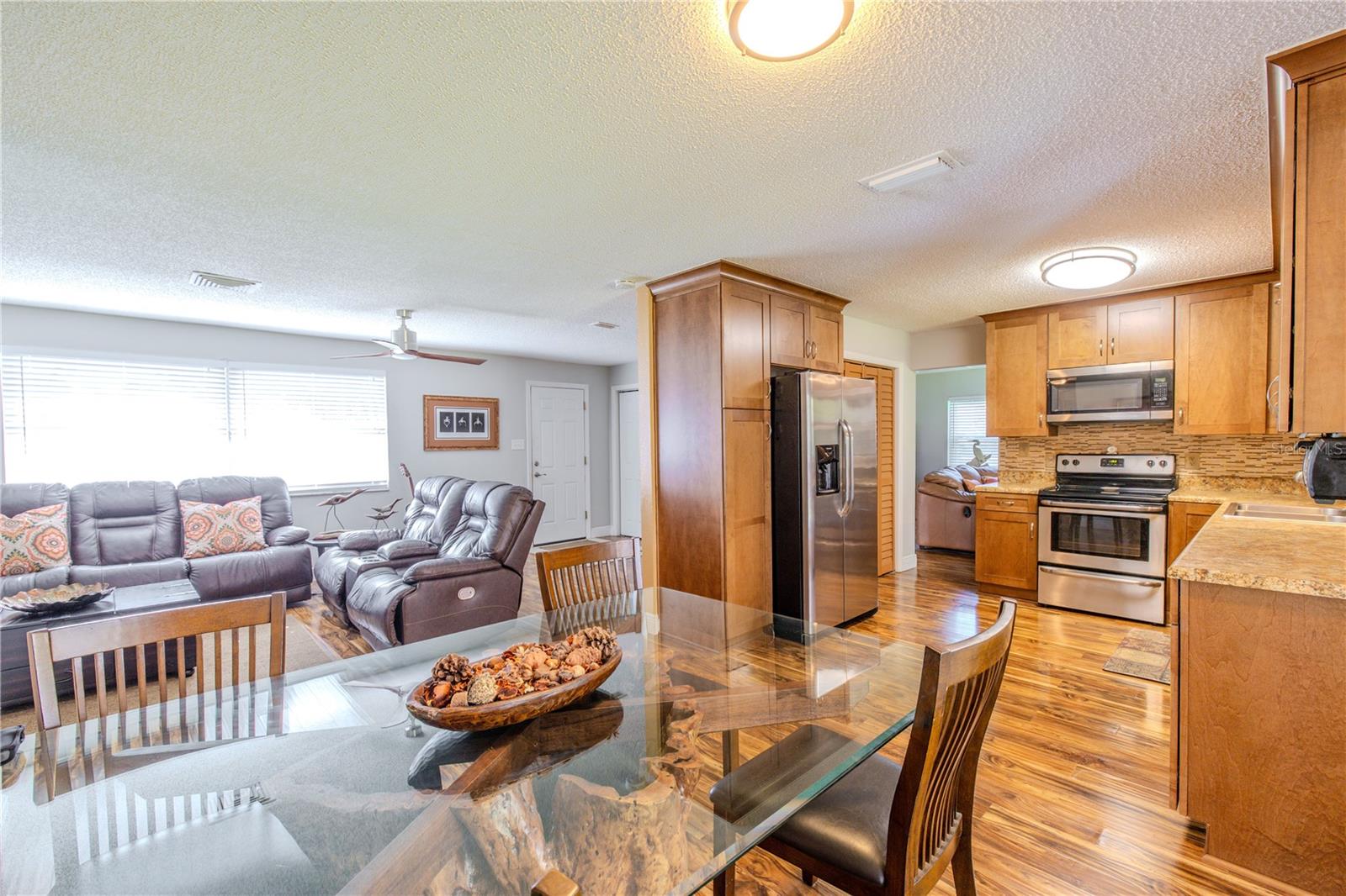 Open view of the living room, dining area, and kitchen, featuring laminate flooring throughout, warm wood cabinetry, and clear sightlines that connect the main living spaces.