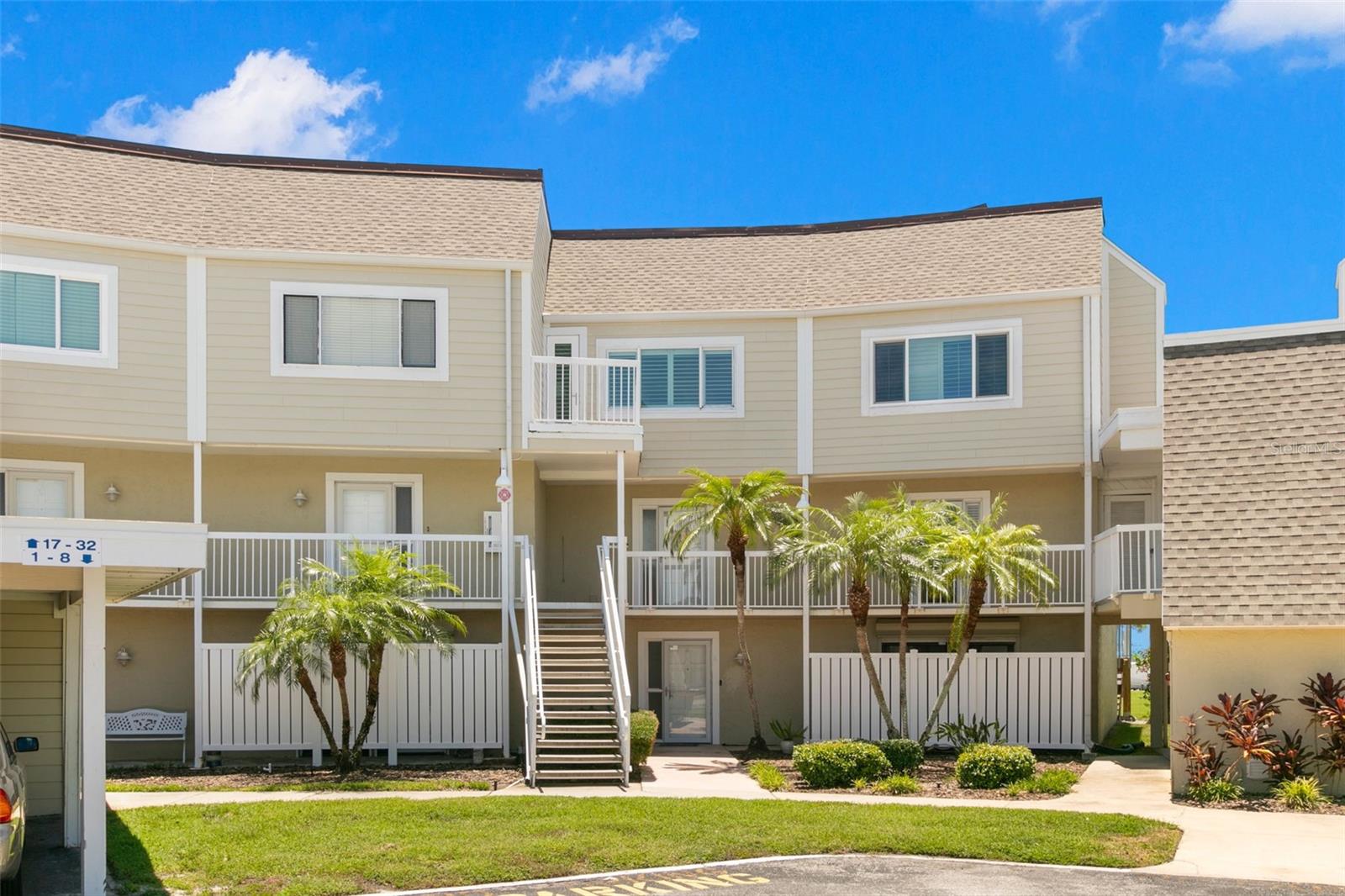 Well-kept coastal-style townhome building with light neutral siding, covered walkways, and a landscaped courtyard entry framed by mature palms.