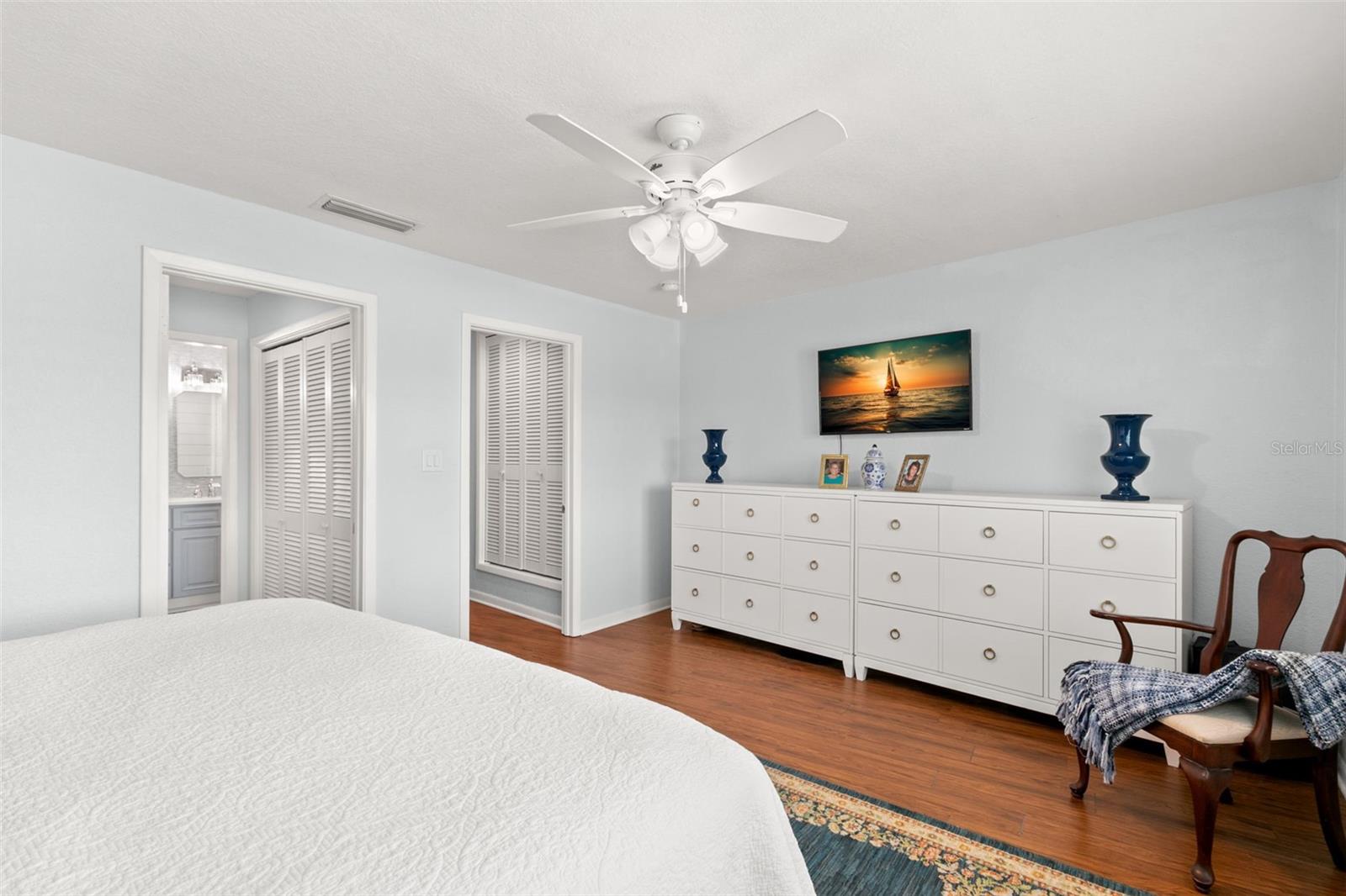Primary bedroom showcasing wood flooring, a ceiling fan, generous wall space, and direct access to the ensuite bathroom and dual closets.