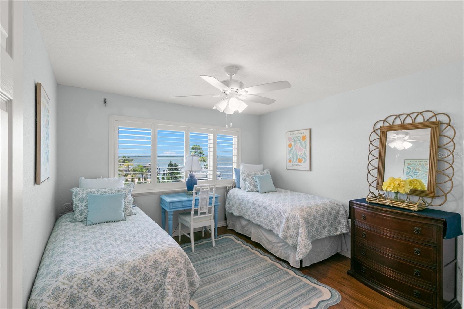 Bright and inviting bedroom featuring luxury vinyl flooring, a ceiling fan, and wide windows fitted with plantation shutters that frame tranquil water views and bring in abundant natural light.