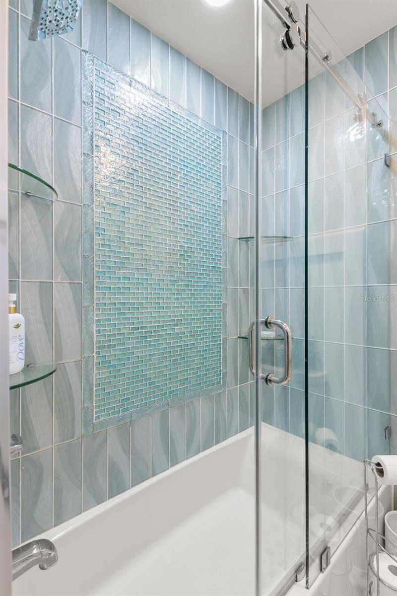 Tub-shower combination with frameless glass enclosure, decorative tile surround, and built-in shelving, offering both style and functionality.