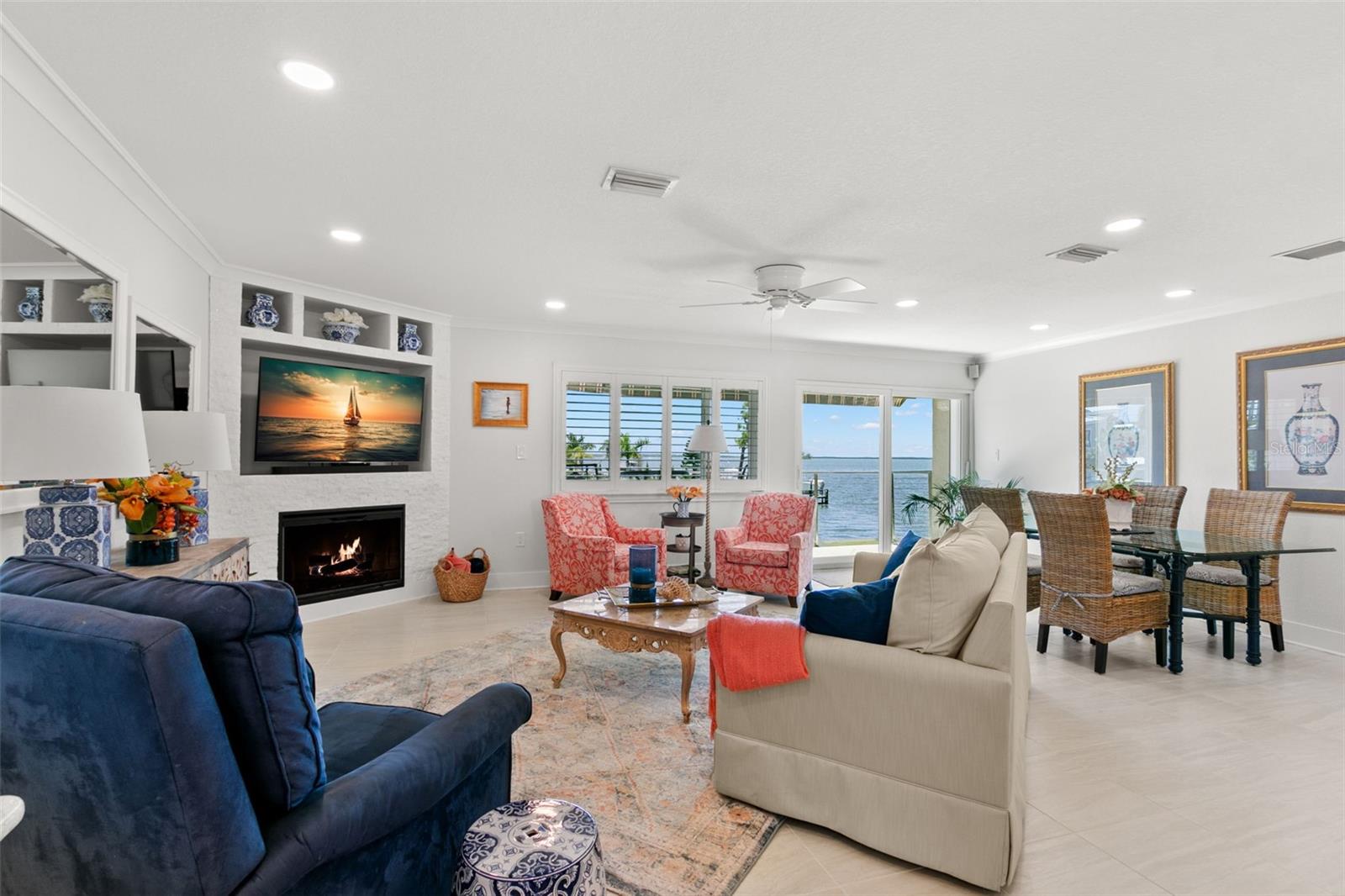 Light-filled living room with recessed lighting, crown molding, and a custom electric fireplace, opening seamlessly to the dining area and sliding doors with expansive water views.