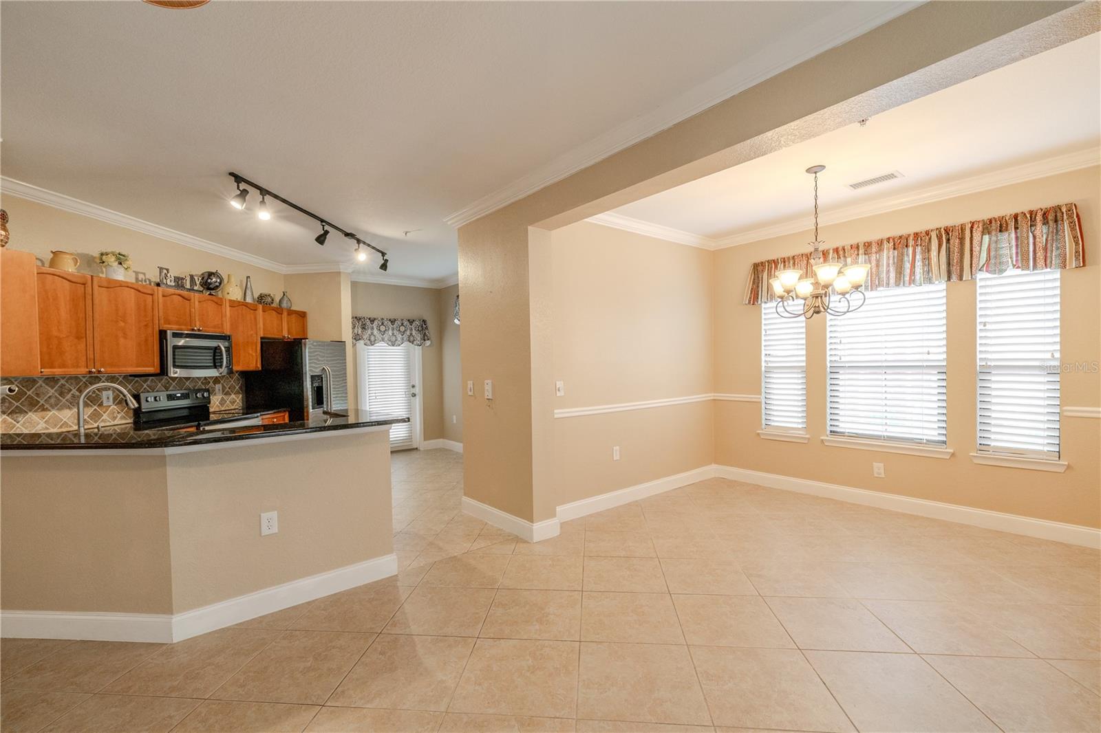 Spacious open-concept living and dining area with neutral tile flooring, crown molding, chair rail detailing, and seamless flow into the kitchen—ideal for everyday living and entertaining.