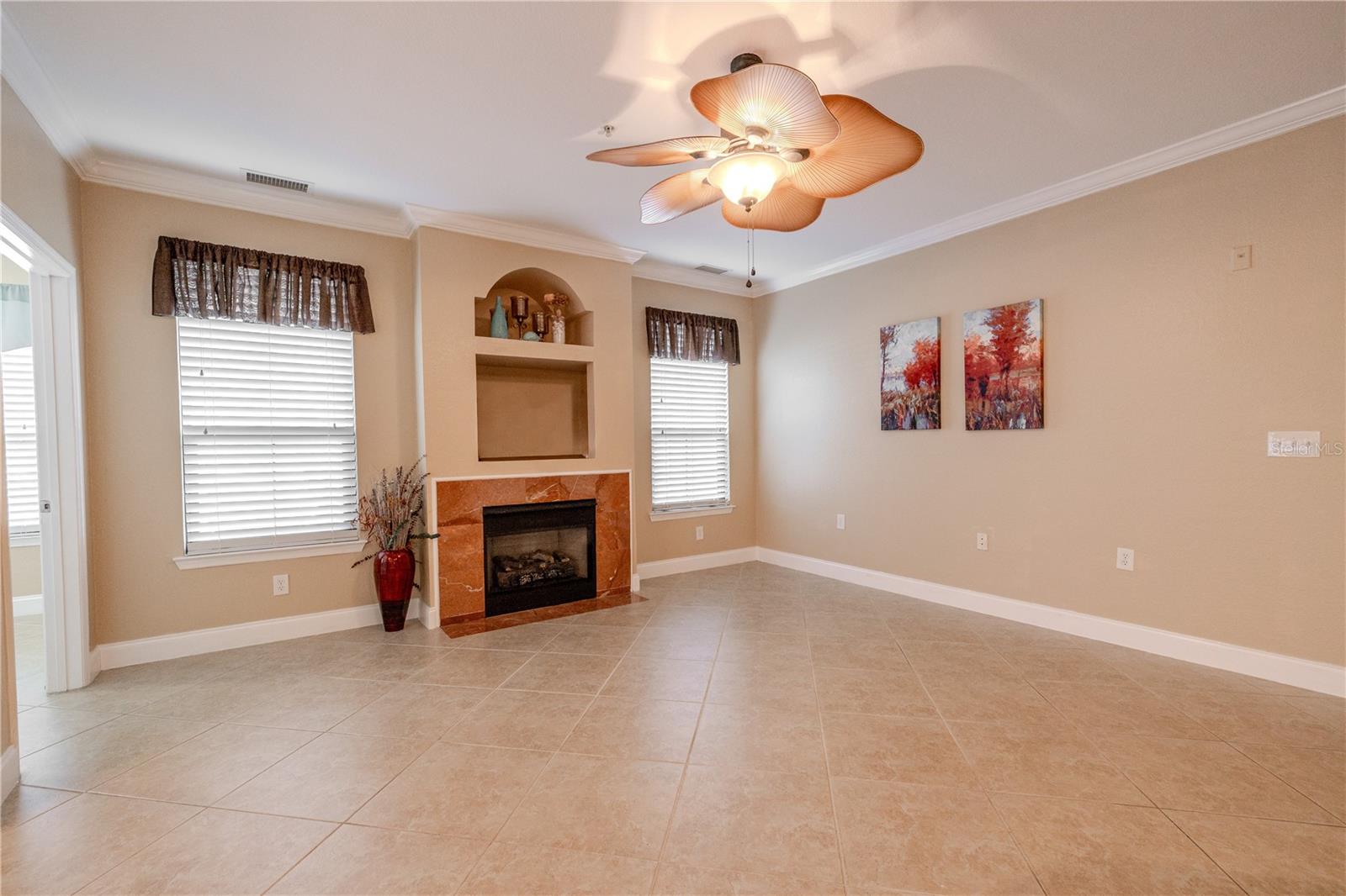 Living room features a tiled fireplace with built-in display niches, crown molding, neutral tile flooring, and a decorative leaf-style ceiling fan, offering a bright, open living space.