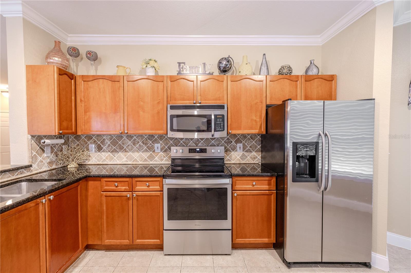 Warm wood cabinetry is paired with granite countertops, a decorative tile backsplash, and stainless steel appliances, including a side-by-side refrigerator and built-in microwave. Crown molding above the cabinets adds a finished, cohesive look to the space.