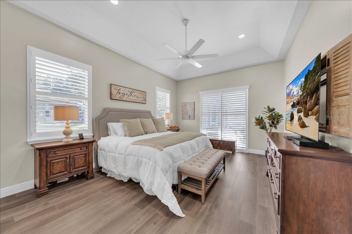 Serene primary bedroom with tray ceiling, ceiling fan, plantation shutters, and warm luxury vinyl flooring!