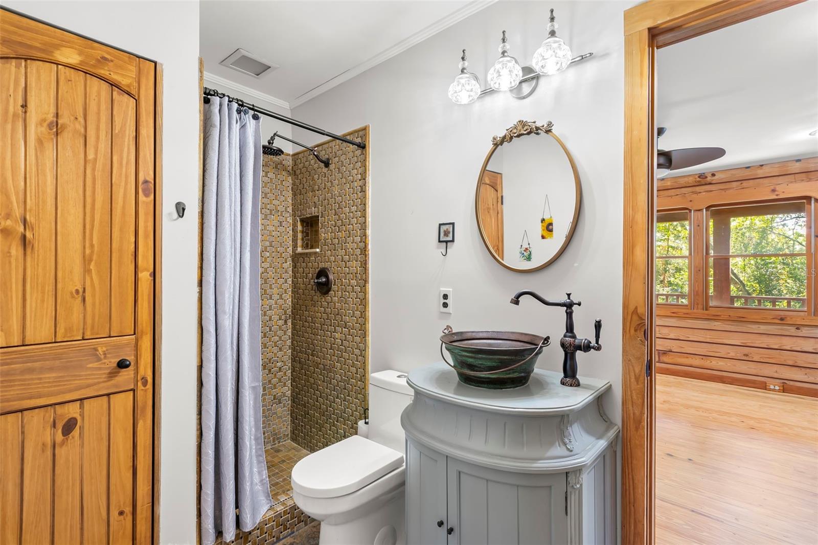 Downstairs bathroom featuring a walk-in tiled shower with niche, furniture-style vanity topped with a vessel sink, oil-rubbed bronze fixtures, crown molding, and a linen closet with a wood door—one of the warm wood accents found throughout the home.