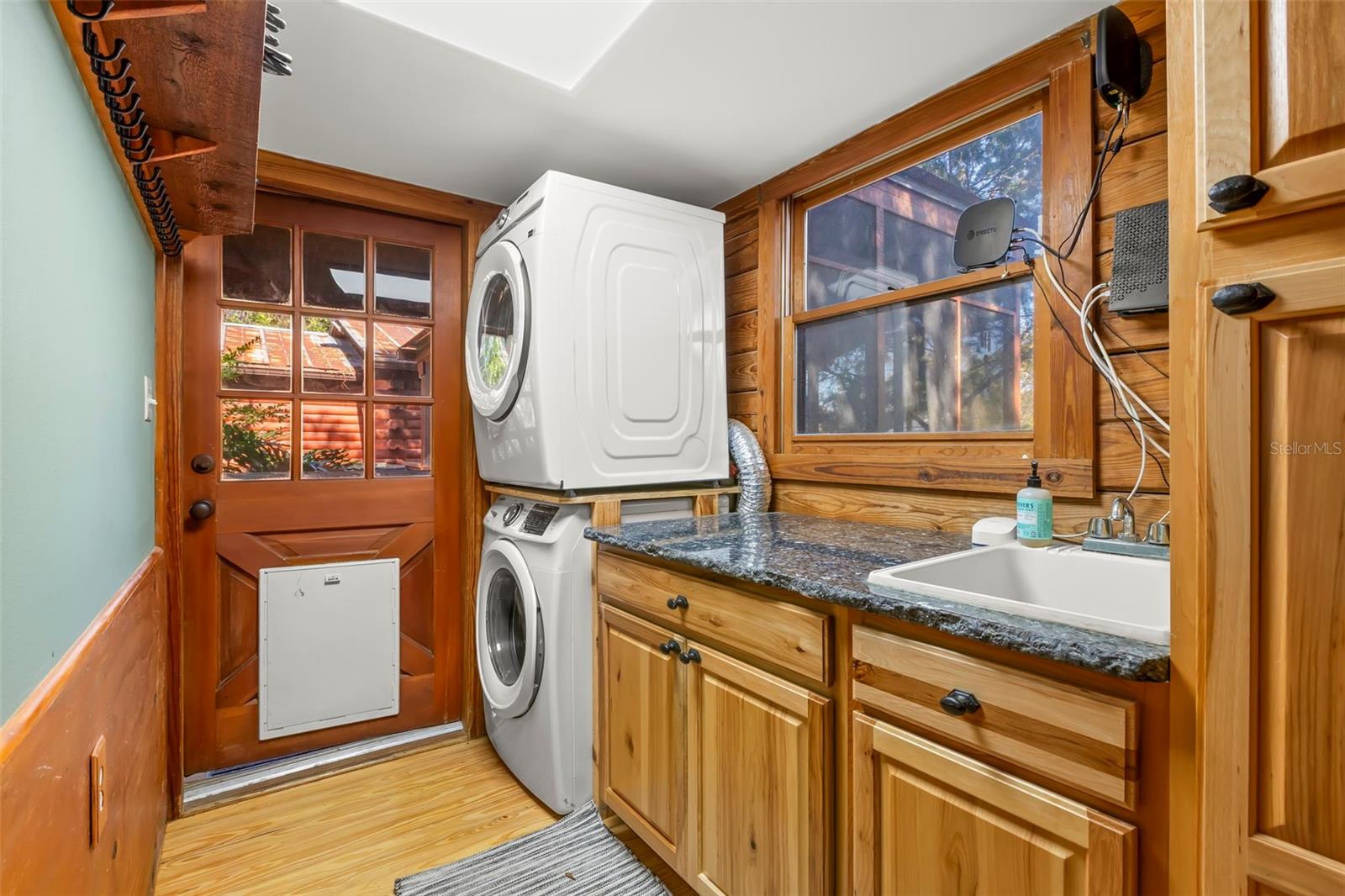 Well-appointed laundry room with full-size front-loading washer and dryer, custom wood cabinetry, granite counters, utility sink, wood flooring, and a glass-paneled wood door—thoughtfully designed to match the home’s natural materials and high-end finishes.