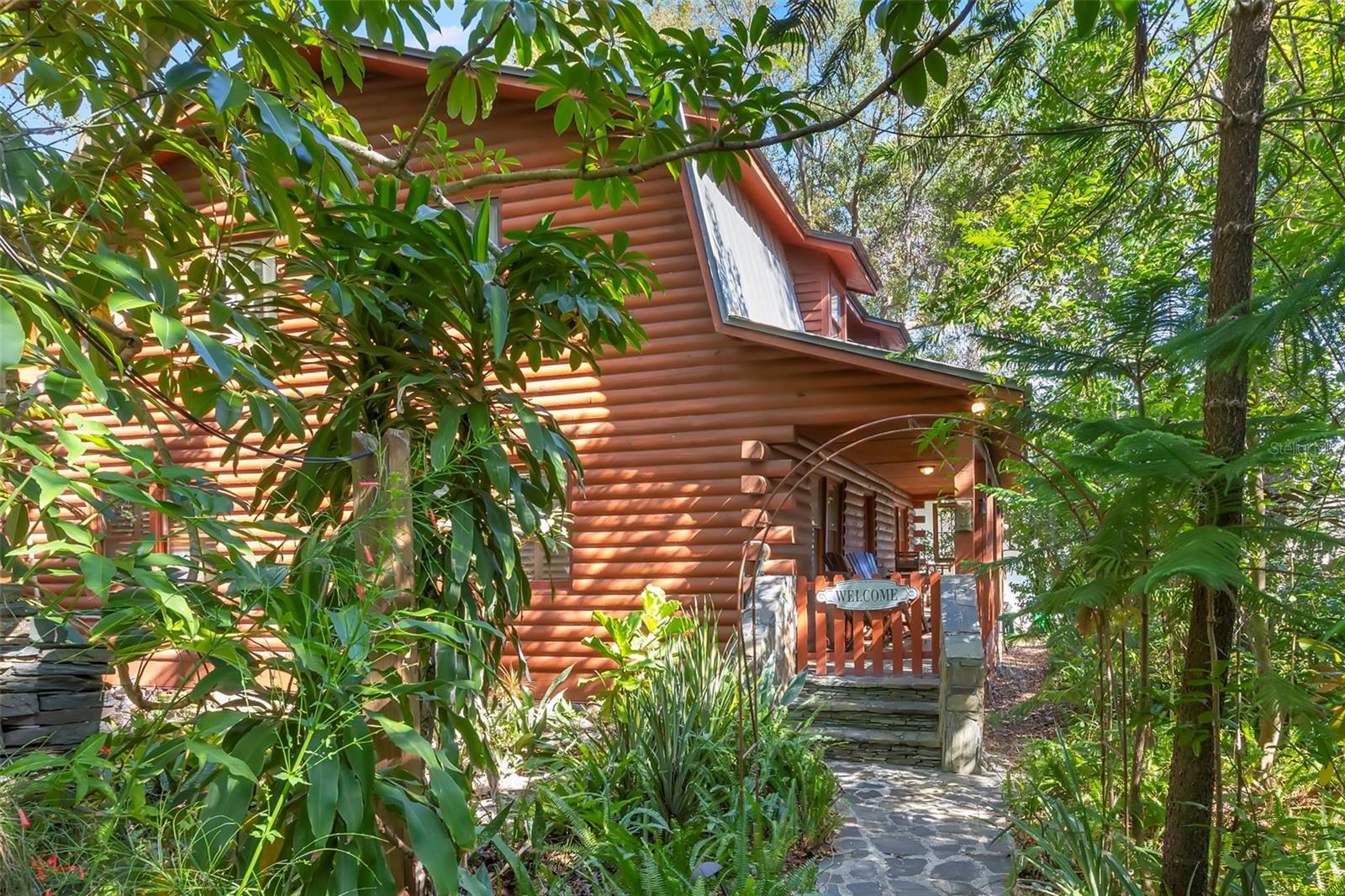 Inviting stone walkway leading to the front porch, framed by lush greenery and a charming gated entry that sets a welcoming first impression. A rare log-style retreat set beneath a canopy of mature trees, offering timeless craftsmanship, deep porch living, and a truly secluded, storybook setting.