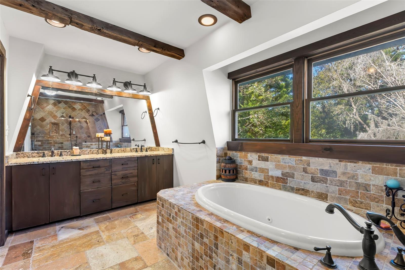 A spa-inspired primary bath anchored by a deep soaking tub set within natural stone tile, framed by wood-trimmed windows and exposed ceiling beams that bring warmth and architectural character to the space.