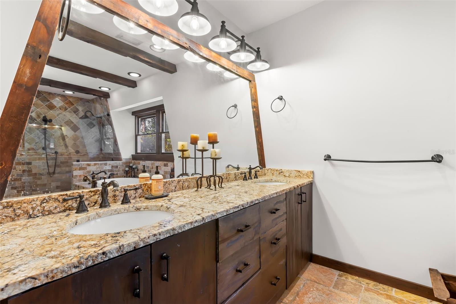 Custom wood cabinetry paired with granite countertops, dual sinks, and oil-rubbed bronze fixtures create a refined vanity area, complemented by a substantial wood-framed mirror and layered lighting for both function and ambiance.