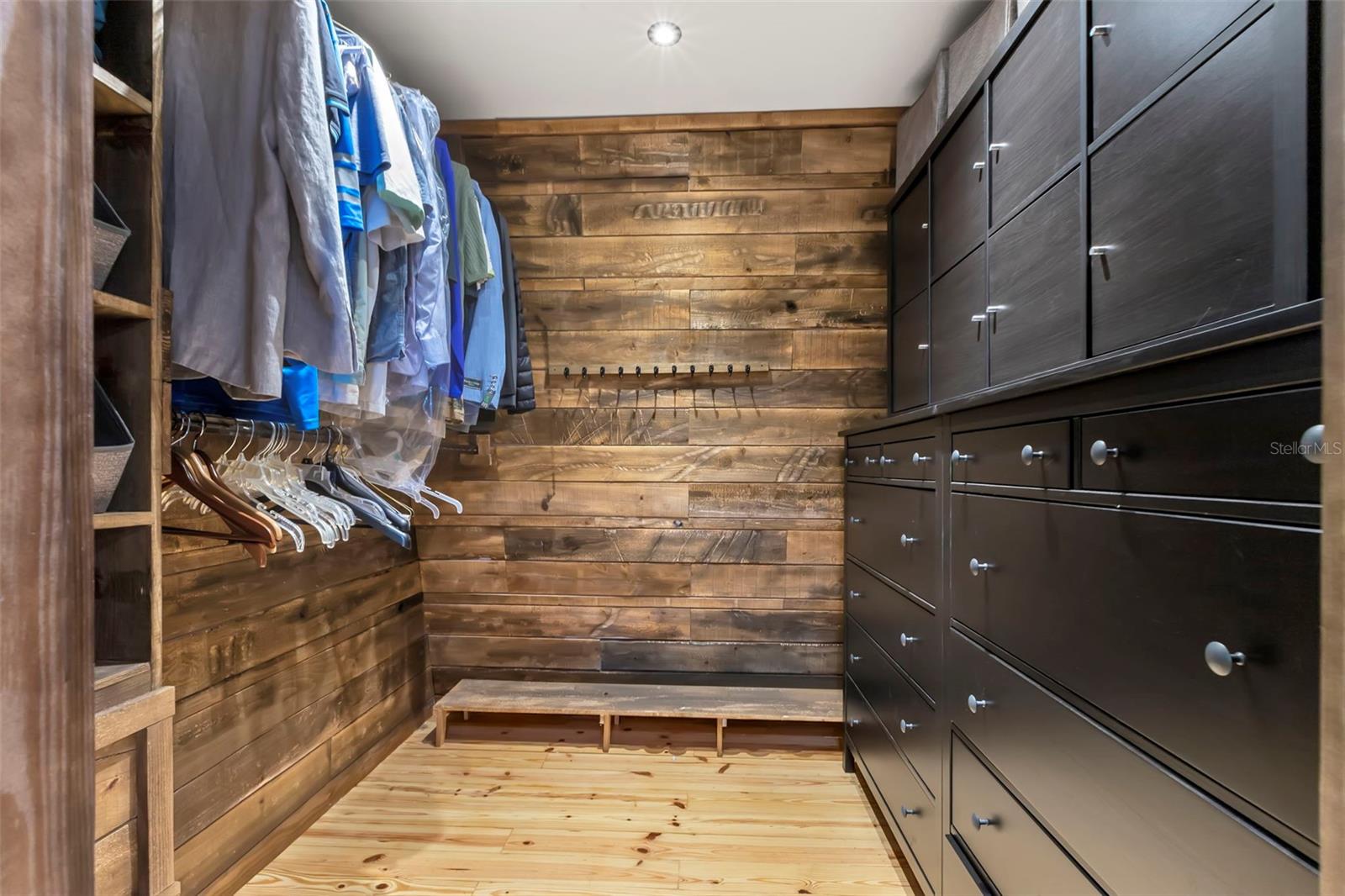 A custom walk-in closet designed as a true dressing room, featuring floor-to-ceiling cabinetry, dedicated hanging space, wood-accent walls, built-in shelving, and a central bench—beautifully balancing storage, organization, and the home’s signature rustic character.
