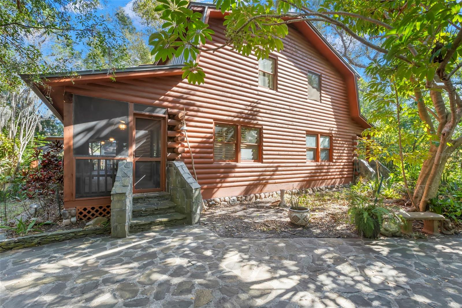 Side view highlighting the authentic log exterior, stone-accented entry steps, and screened porch addition tucked into the natural surroundings.