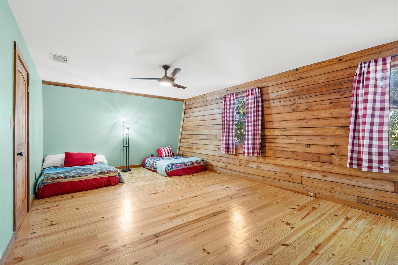 This oversized upstairs bedroom offers a warm, lodge-inspired atmosphere with beautifully finished wood floors and rich natural textures throughout. The sloped ceiling adds architectural interest, while solid wood doors and trim reinforce the home’s thoughtful attention to detail. A contemporary ceiling fan provides quiet comfort, and the open layout allows for versatile furnishing options—ideal for guests, a shared sleeping space, or a creative retreat.