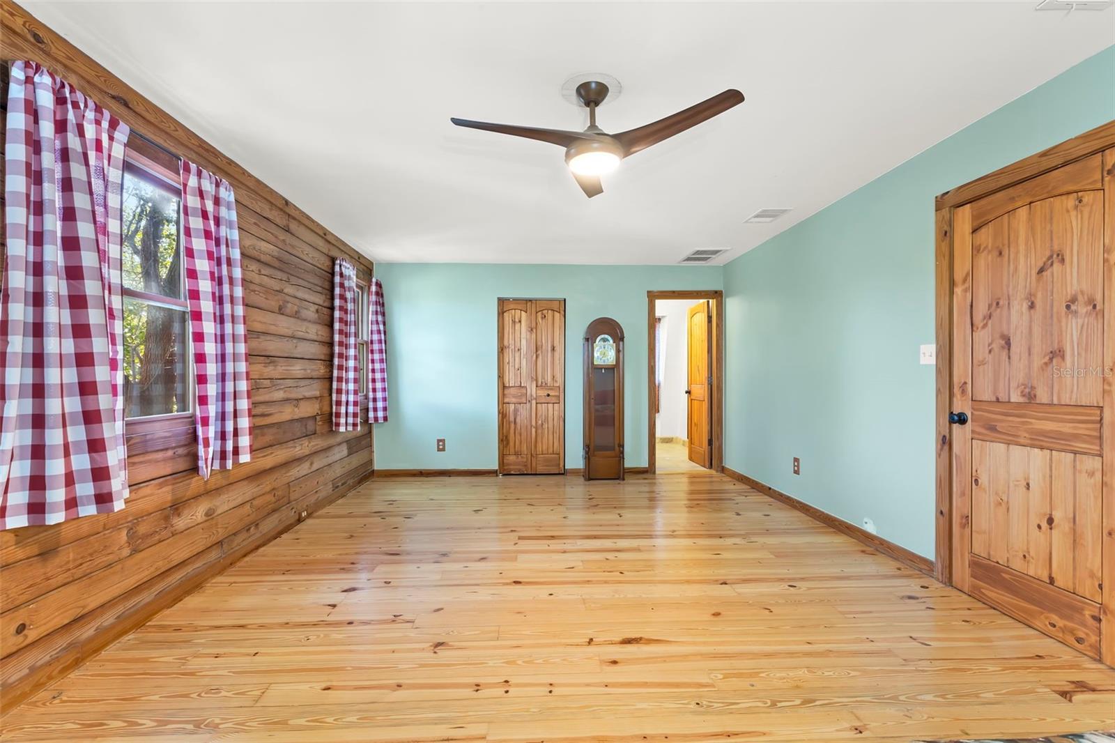 Tucked away on the upper level, this expansive bedroom blends rustic character with airy comfort. Wide-plank wood floors, a wood-paneled accent wall, and soft coastal-toned walls create a relaxed retreat with timeless charm. A modern ceiling fan adds year-round comfort, while multiple wood-trimmed windows and solid wood doors echo the home’s cohesive craftsmanship. The generous footprint easily accommodates multiple beds, a seating area, or flexible use as a guest suite, studio, or secondary primary-style bedroom.