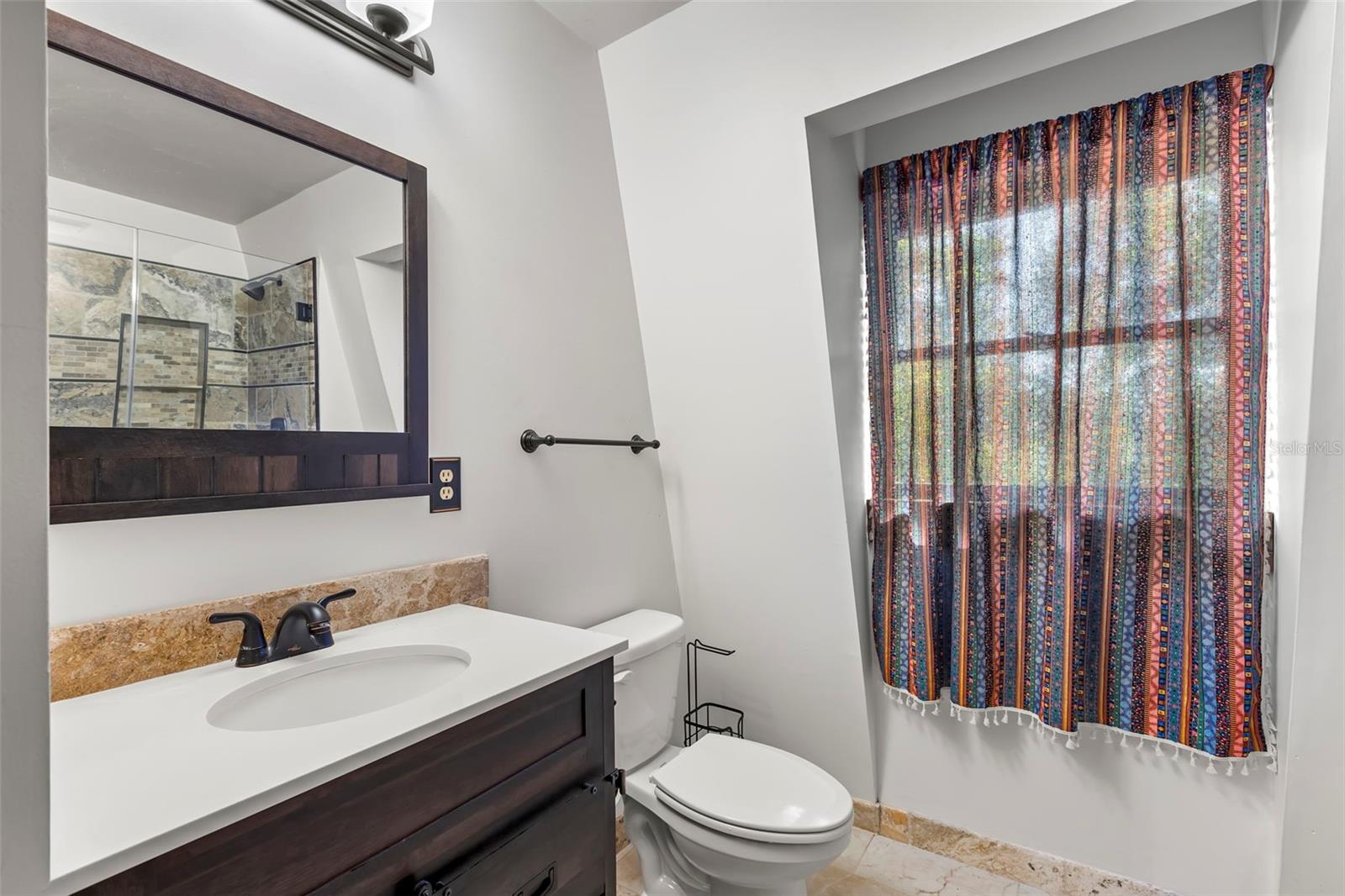 A thoughtfully designed 3rd bedroom ensuite bath pairs clean architectural lines with warm materials, featuring a rich wood vanity topped with a smooth, solid-surface countertop and an integrated sink. Dark hardware and fixtures add contrast, while a framed mirror and contemporary wall lighting create a polished, comfortable space for daily routines. A deep-set window alcove brings in soft natural light and adds subtle character without sacrificing privacy.