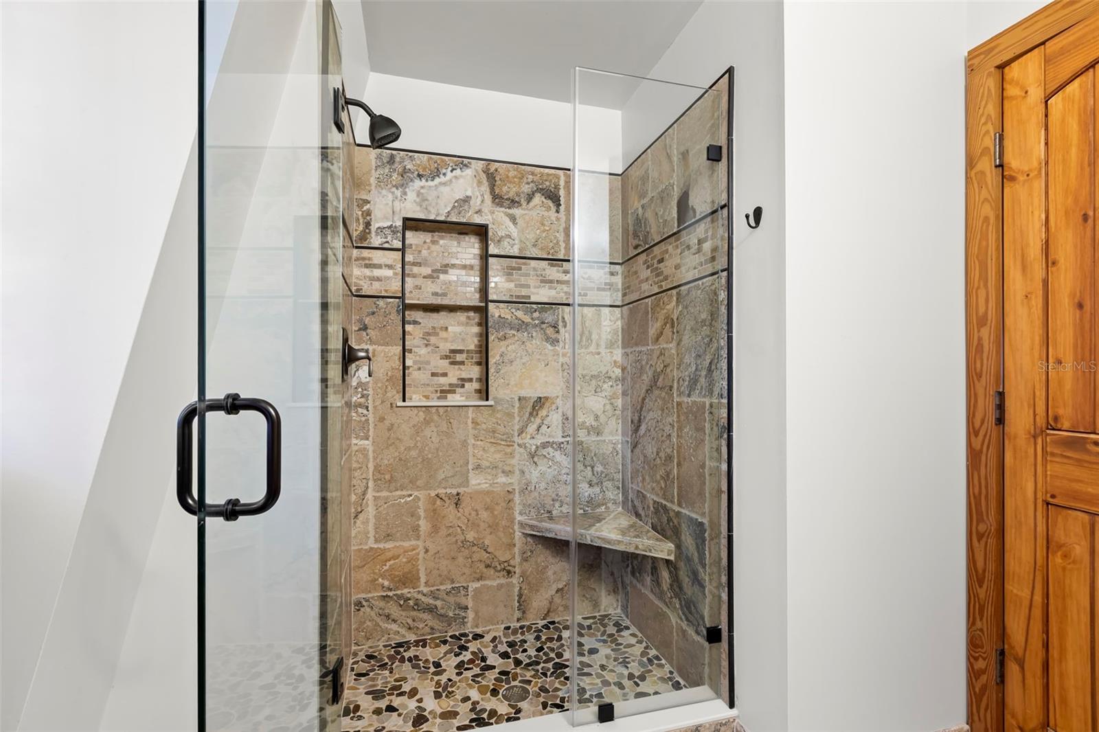 This 3rd bedroom ensuite walk-in shower delivers a spa-inspired experience with floor-to-ceiling natural stone tile, a frameless glass enclosure, and a striking pebble-style shower floor underfoot. A built-in corner bench and recessed niche add both function and design, while dark fixtures provide a modern counterpoint to the warm stone palette. Clean lines, quality finishes, and thoughtful details elevate this private bath beyond the expected.