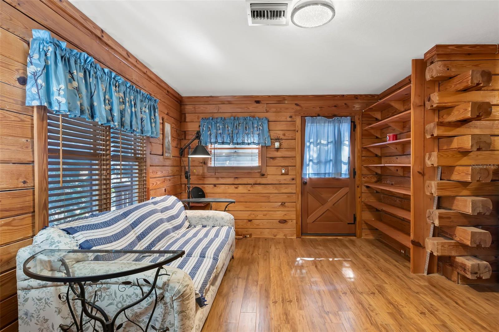 Warm, lodge-style character of the guest house defines this living space, where horizontal solid-wood plank walls are accented by exposed log ends that add authentic cabin craftsmanship. Built-in open shelving is seamlessly integrated into the log structure, offering both display and storage while reinforcing the home’s rustic, handcrafted aesthetic. Natural wood floors and stone-accent walls complete the cozy, retreat-like atmosphere.