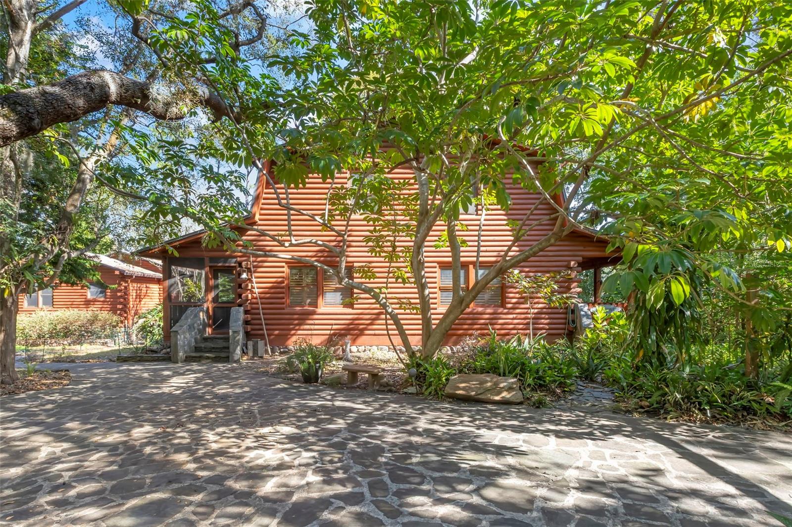 Wide view of the stone-paved drive and courtyard area, offering a serene approach to the home with abundant shade and natural texture.