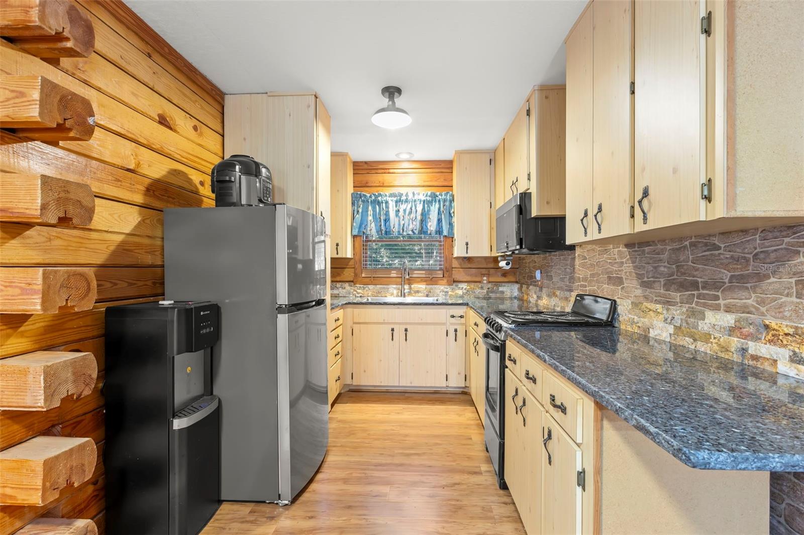 The guest house kitchen is framed by solid-wood log walls with visible log ends that highlight the home’s authentic construction and craftsmanship. Light wood cabinetry complements the natural tones, while stone-look backsplash and dark countertops provide contrast and durability. The space balances rustic charm with practical functionality, creating a kitchen that feels both inviting and purposeful.