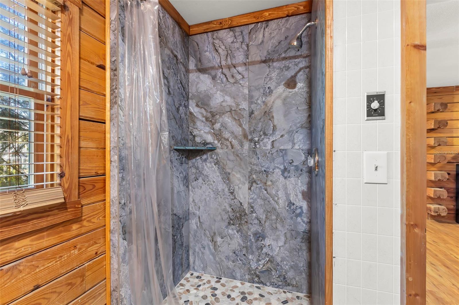 A stone-look shower surround and pebble-style flooring introduce spa-inspired texture, while wood cabinetry and trim maintain the home’s cohesive, natural aesthetic.