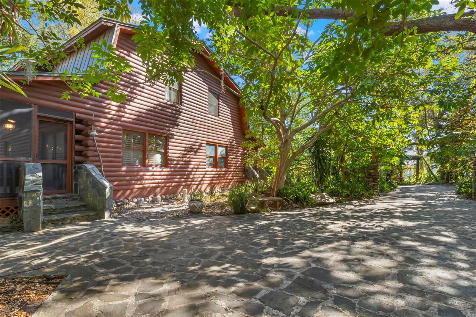 A gently curving stone driveway winds beneath a canopy of mature trees, creating a shaded, welcoming approach to the home. Dappled sunlight filters through the foliage, highlighting the natural textures of the stone and wood surroundings and setting the tone for the private, retreat-like setting beyond.