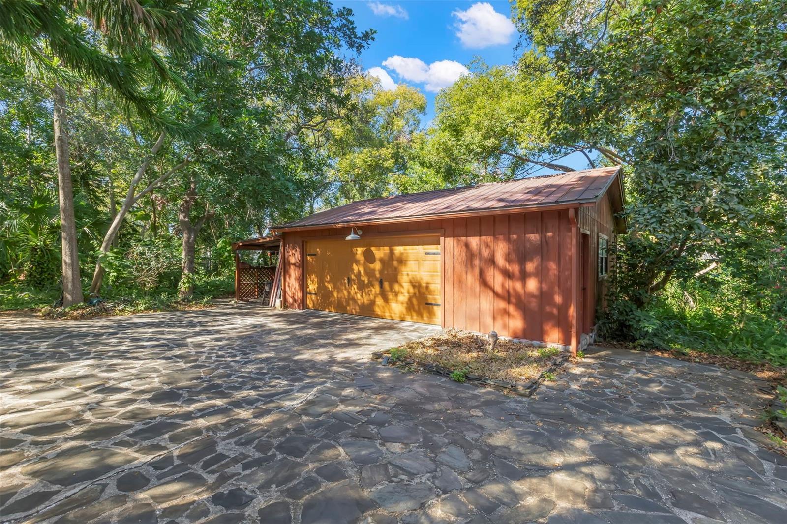 A spacious 2.5-car detached garage featuring a wide wood door with electric opener, generous interior capacity for vehicles, storage, or a workshop, and a stone-paved approach that complements the property’s natural setting. Surrounded by mature trees, the structure blends seamlessly into the landscape while offering practical, everyday functionality.