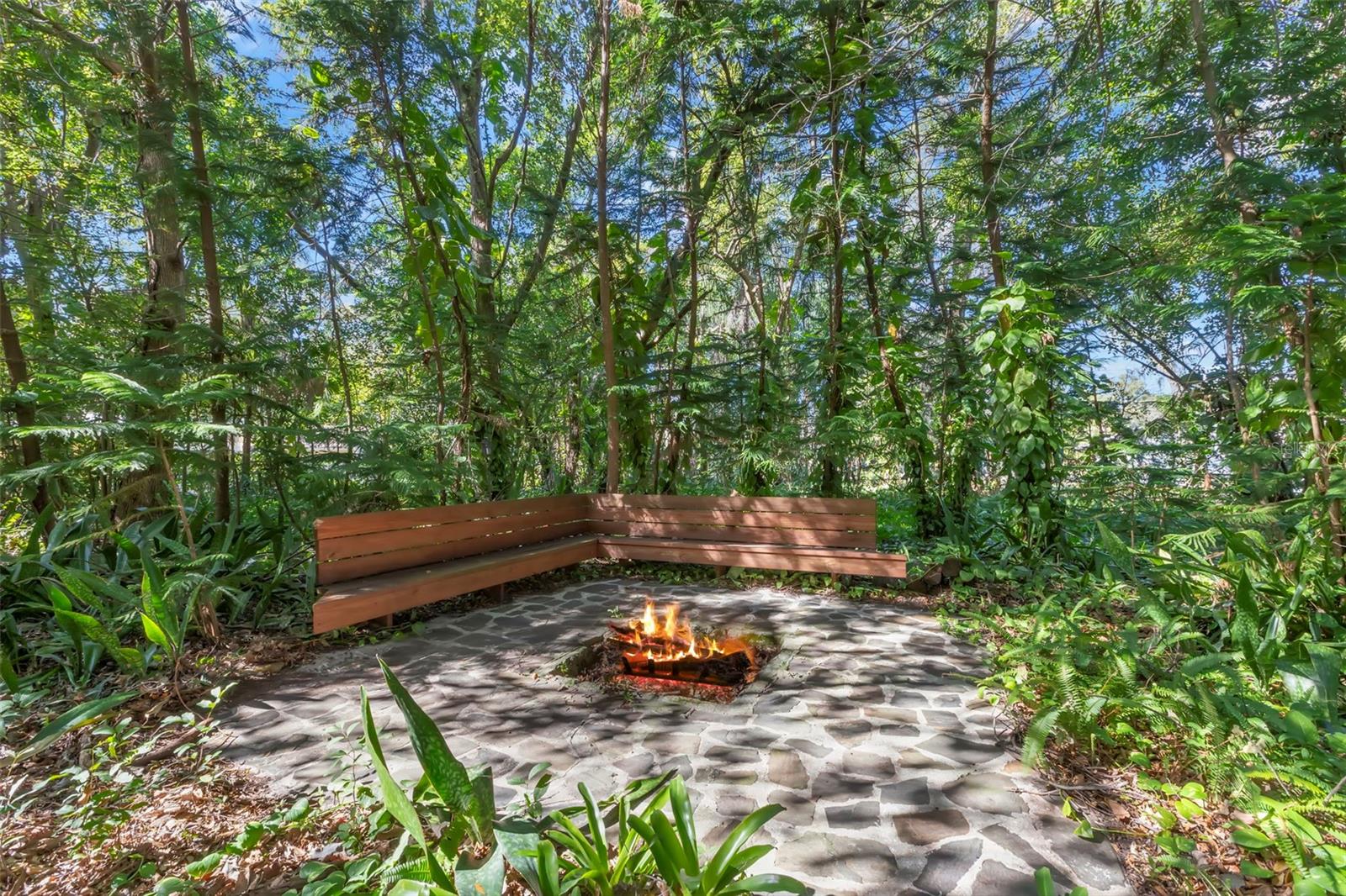 Tucked beneath a canopy of mature trees, this stone-paved fire pit creates a quiet gathering place surrounded by natural greenery. Built-in wood bench seating wraps the space, offering a peaceful setting for evening fires, conversation, and moments of calm that feel far removed from the everyday.