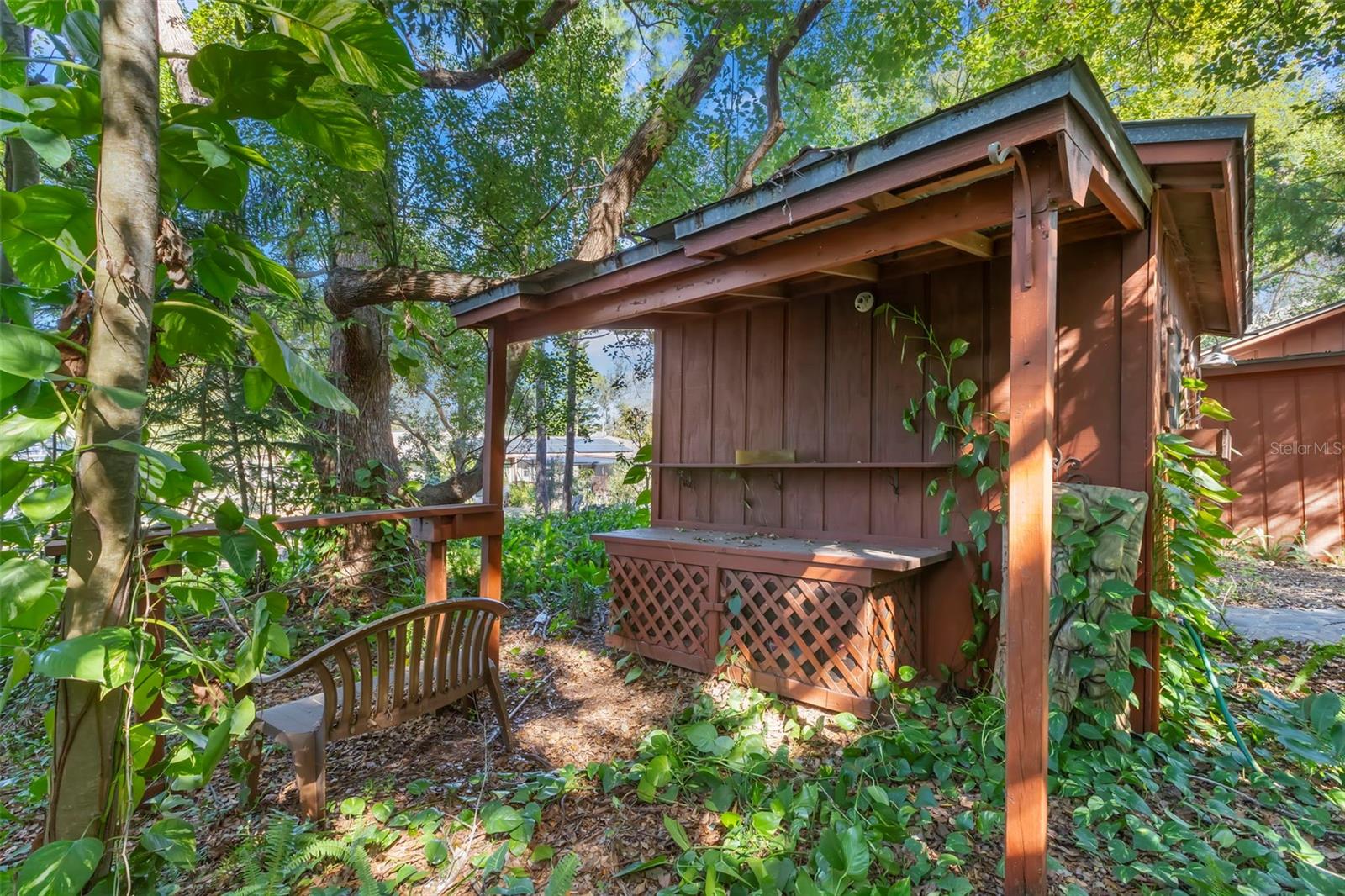 Nestled beneath a leafy canopy, this covered outdoor nook feels like a hidden woodland escape. Framed by mature trees and climbing greenery, the sheltered structure offers built-in shelving, a lattice-front storage bench, and a peaceful spot to sit, read, or simply enjoy the quiet rhythm of the landscape—an intimate outdoor space that feels thoughtfully integrated into its natural surroundings.