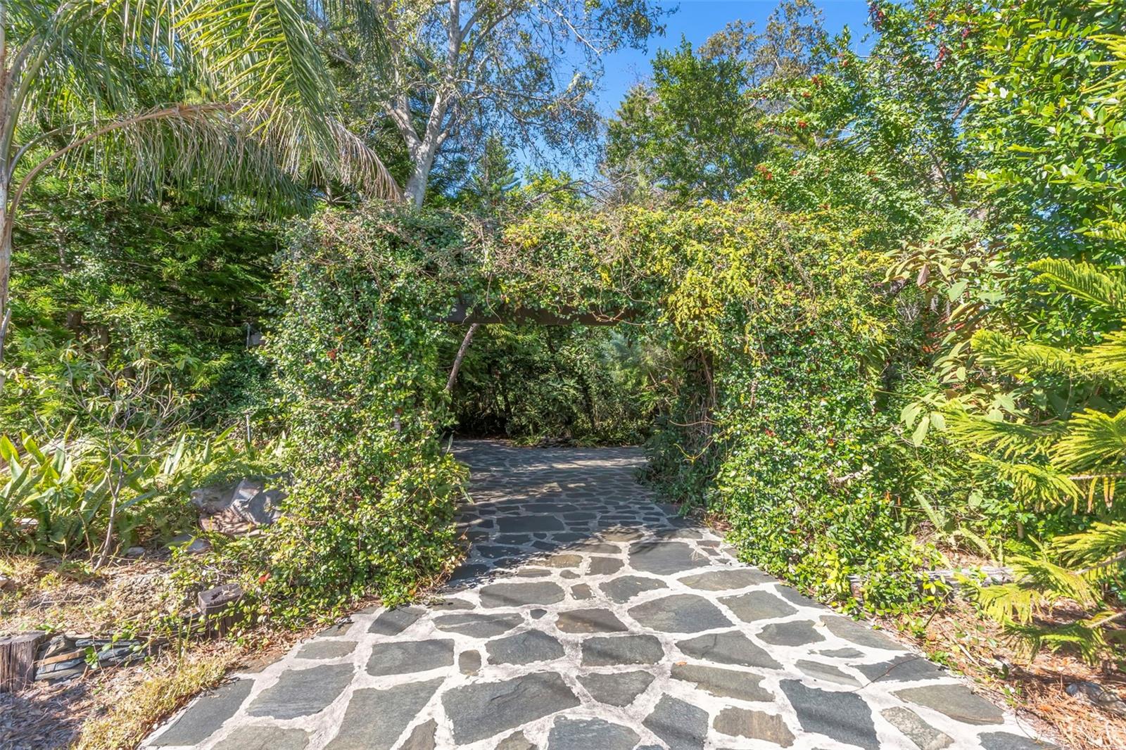 A flagstone-paved driveway leads through a lush, jasmine-covered pergola, creating a fragrant, garden-like arrival framed by mature trees and natural landscaping for privacy and charm.