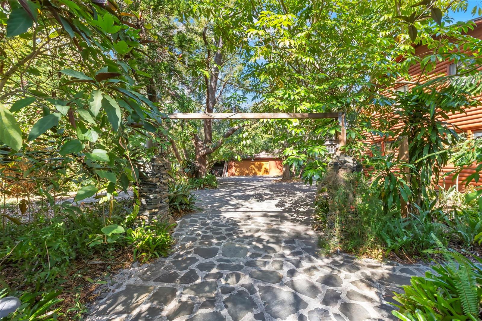 A winding stone driveway passes beneath a rustic timber pergola and mature canopy, creating a shaded, storybook approach framed by lush plantings and natural stone pillars.