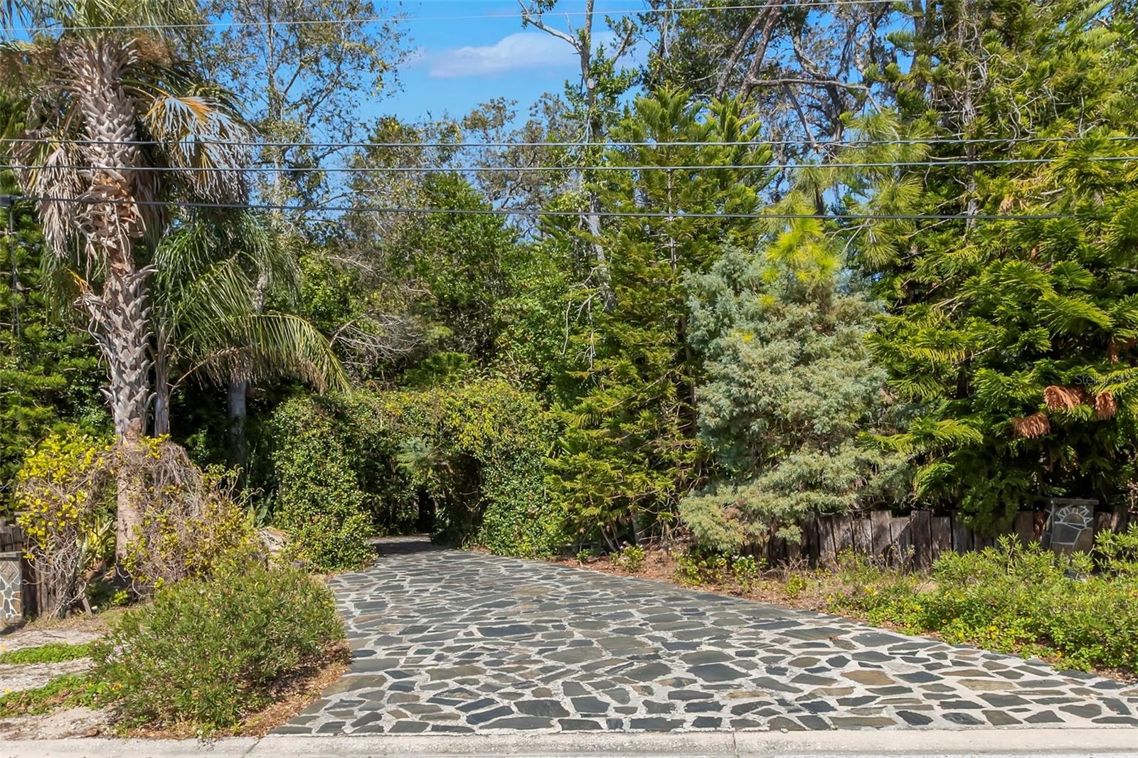 A flagstone-paved driveway leads through a lush, jasmine-covered pergola, creating a fragrant, garden-like arrival framed by mature trees and natural landscaping for privacy and charm.