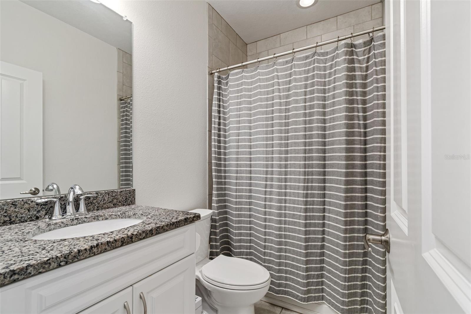 Upstairs full bath with granite countertop, white vanity, and tub/shower combo framed by neutral tile and striped curtain.