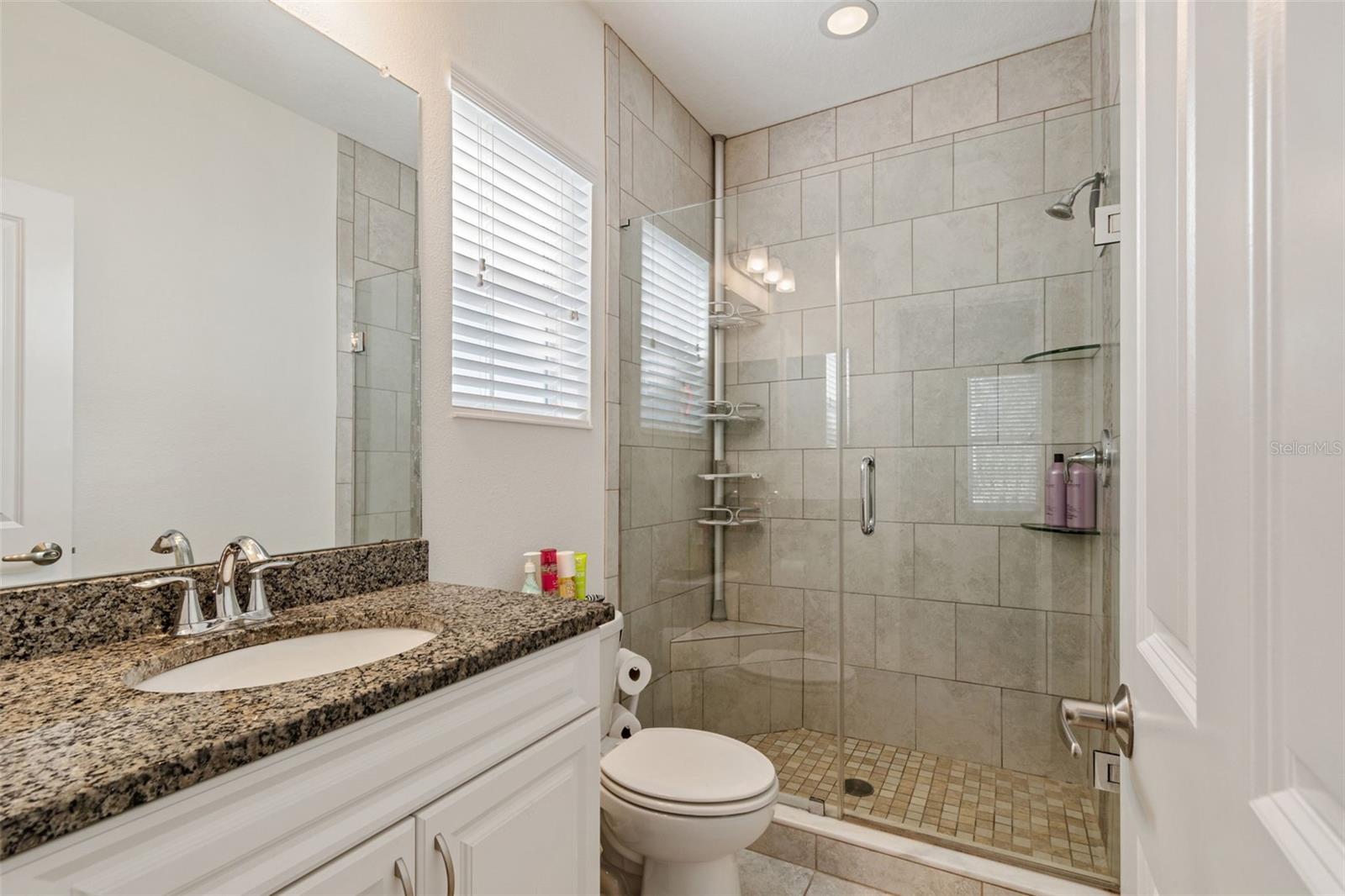 Features a granite vanity, tile floors, and a frameless glass shower with built-in shelving and natural light from a privacy window.