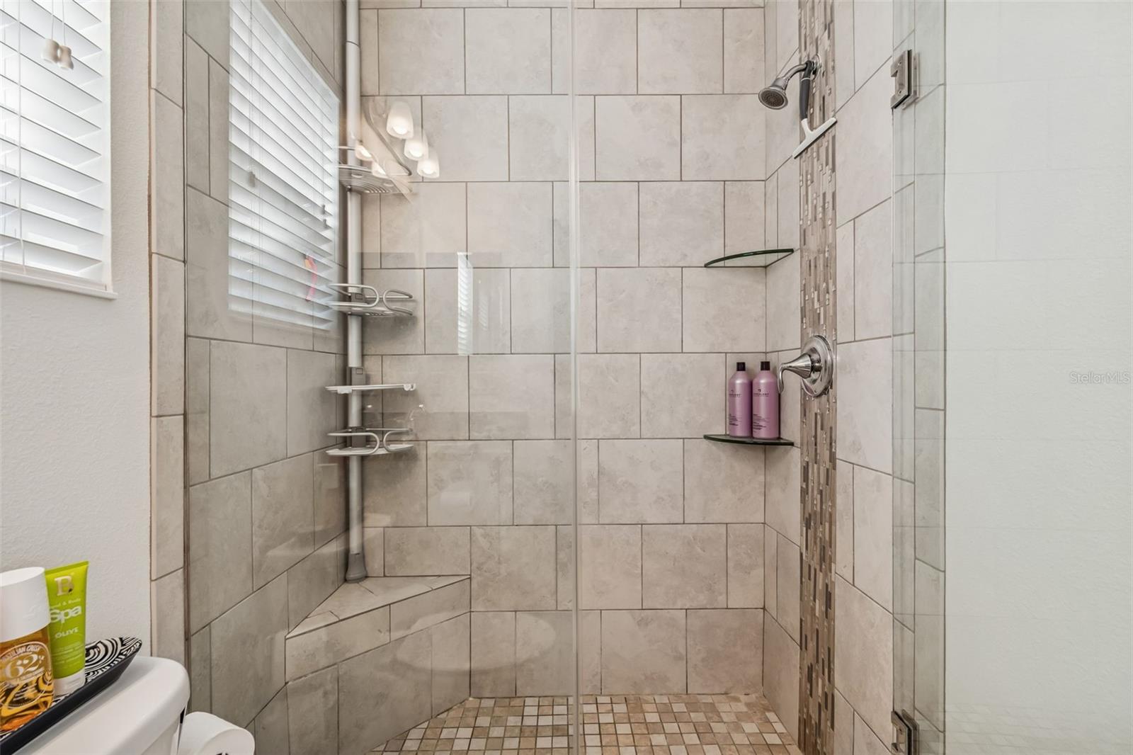 Tiled shower with built-in seat, vertical accent tile strip, and dual corner glass shelves—designed for everyday ease and comfort.